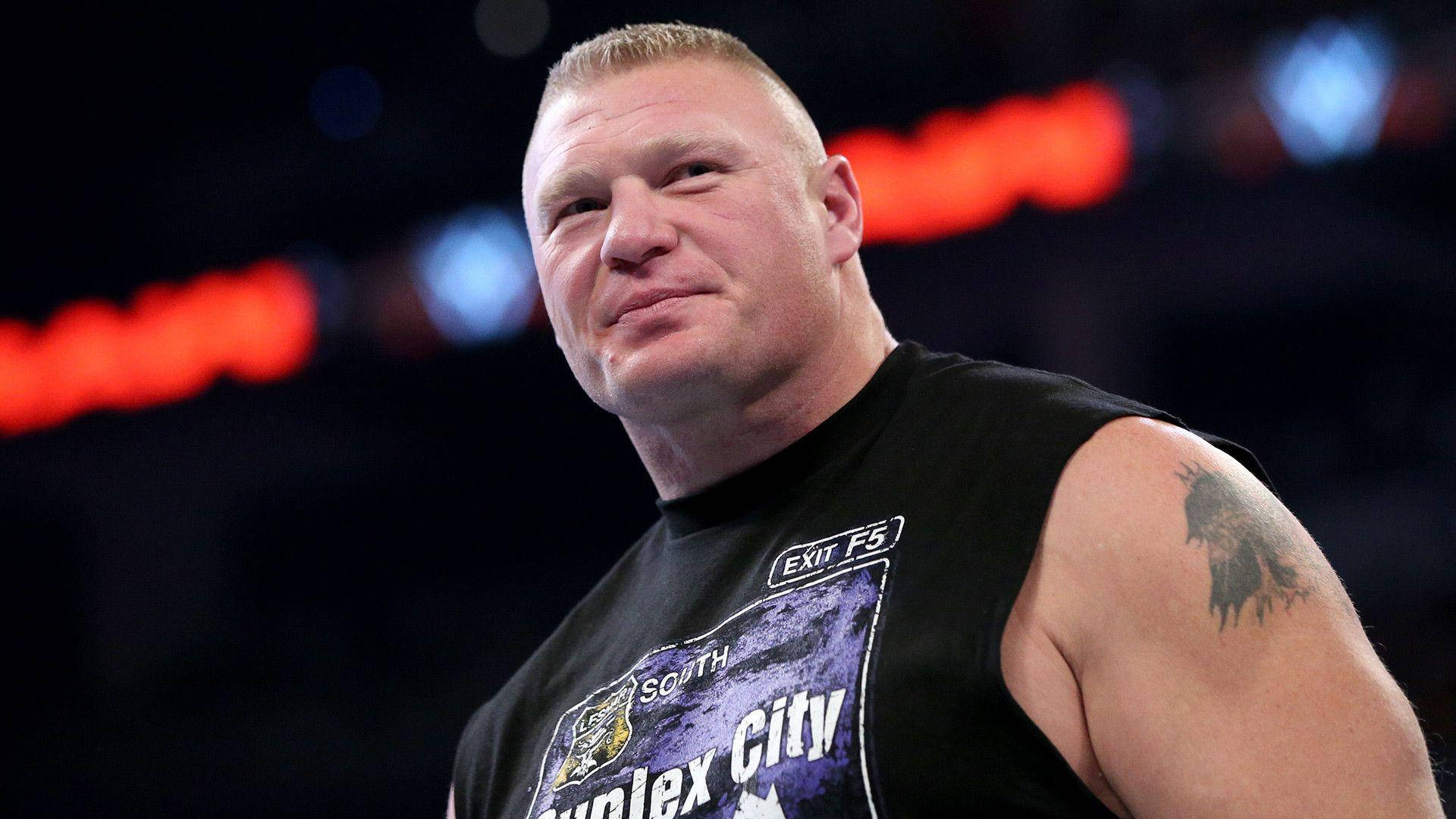 筆電 桌布 壁紙 — 摔角手布洛克·雷斯納（Brock Lesnar）身穿黑色無袖衫，露出刺青手臂，背景是模糊的競技場。