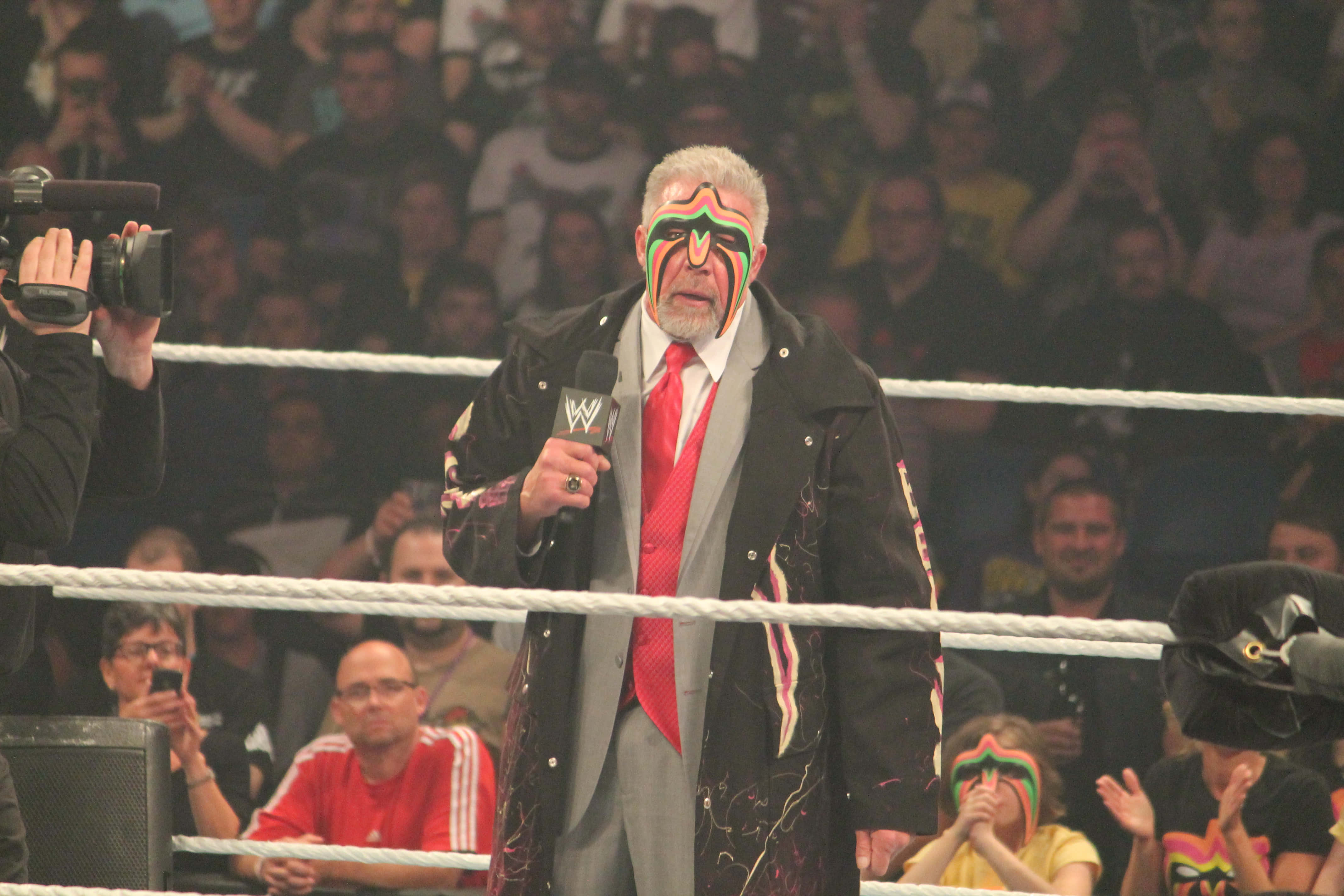 WWE 桌布 壁紙 — 摔角手臉上塗著彩色顏料，站在擂台上拿著麥克風，觀眾熱切觀看。