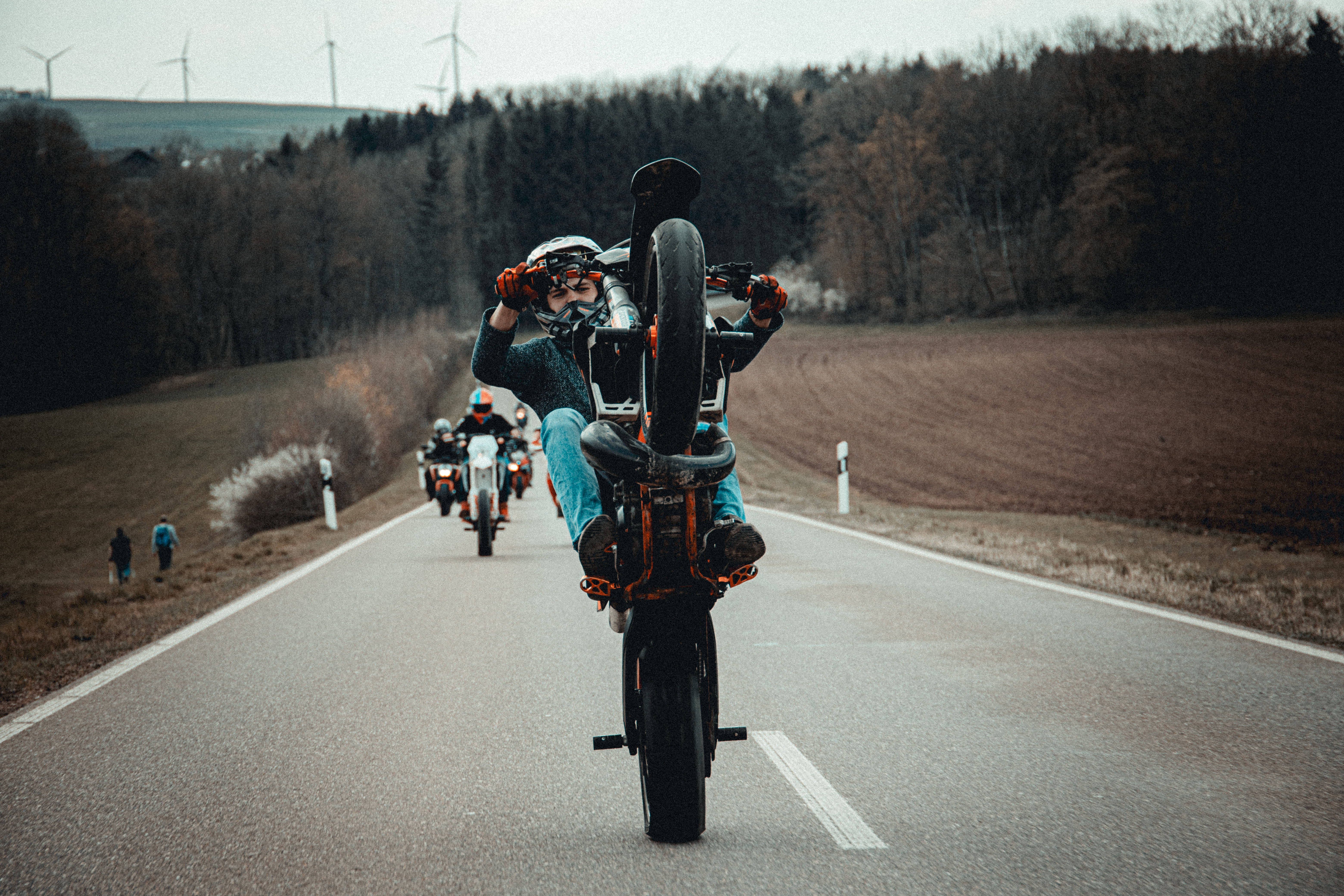 Ktm Bike 桌布 壁紙 — 摩托車手在鄉村道路上表演前輪離地，背景有風力發電機和光禿禿的樹木。