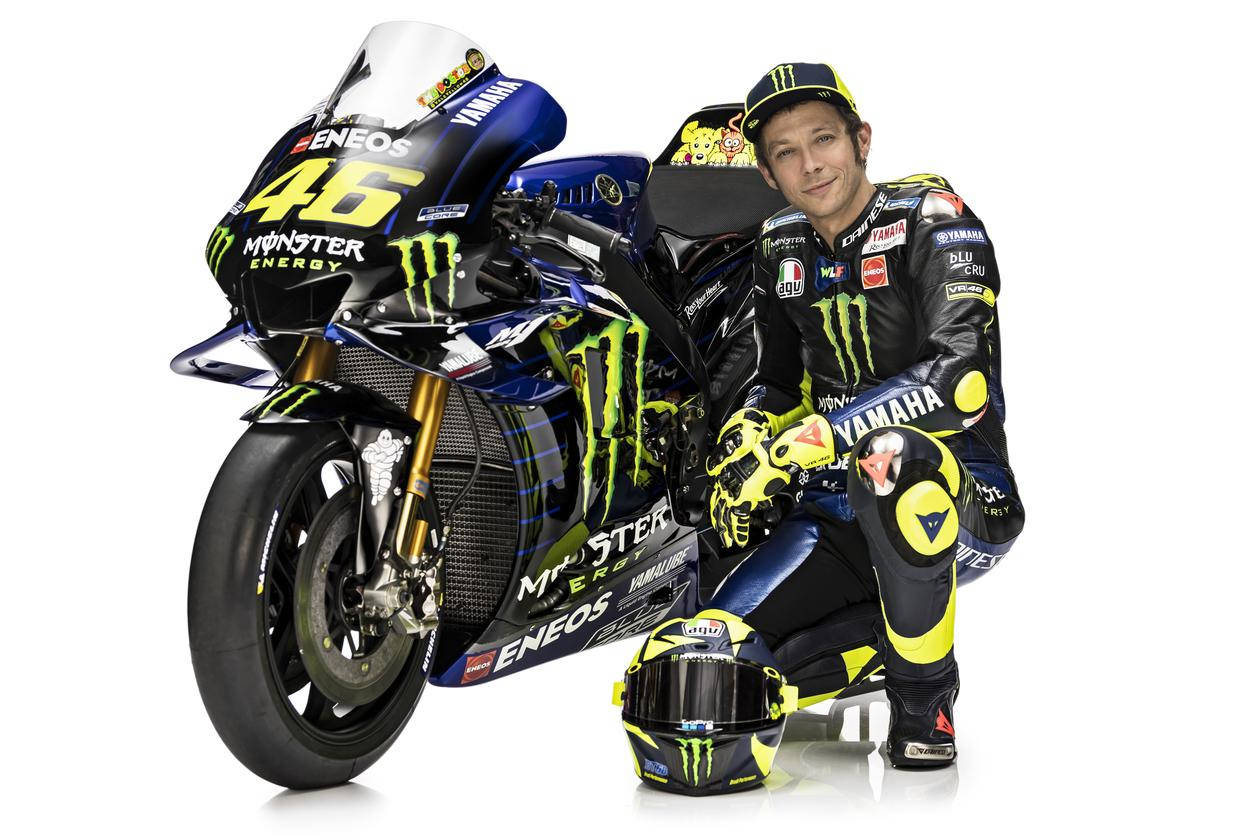 Monster Energy 桌布 壁紙 — 摩托車賽車手瓦倫蒂諾·羅西身穿全套裝備，跪在他藍黃色的Yamaha MotoGP摩托車旁，背景為白色。