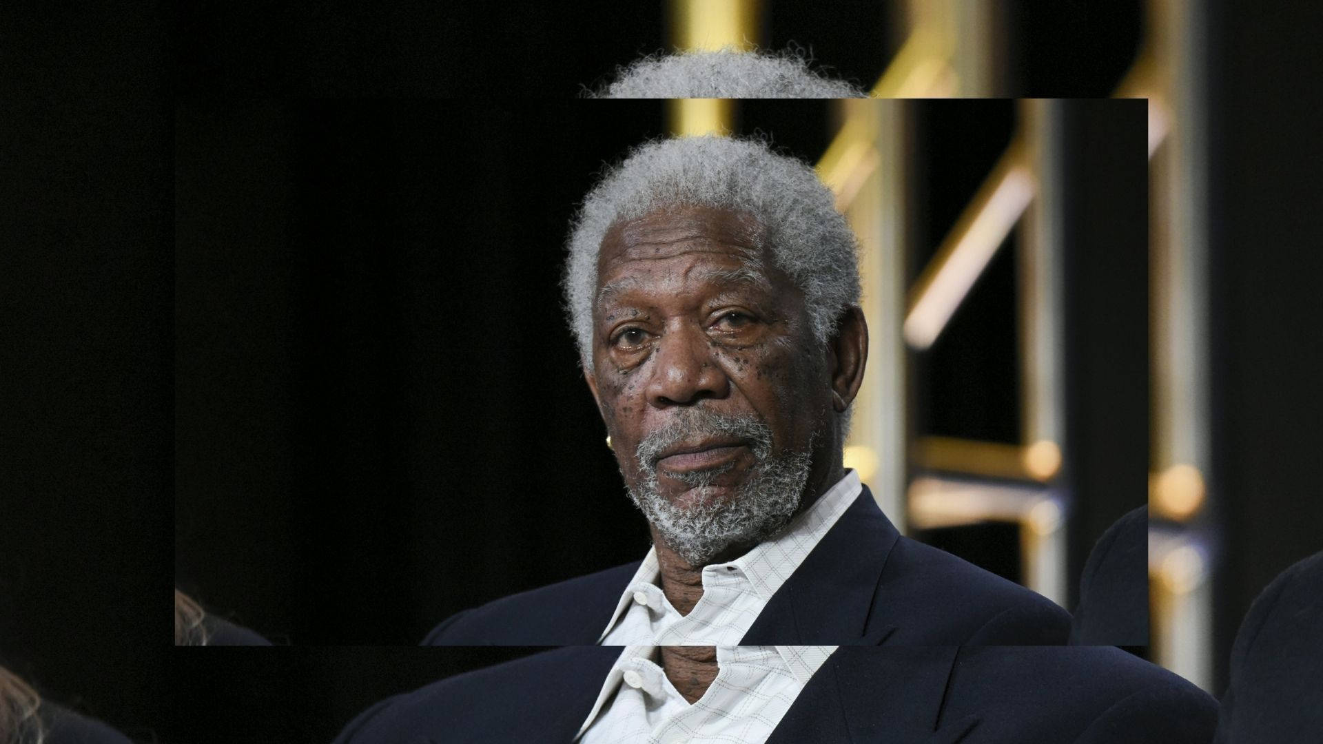 Morgan Freeman 桌布 壁紙 — 摩根費曼的頭像近照，灰髮灰鬚，身穿深色西裝，背景昏暗，金色舞台燈光。