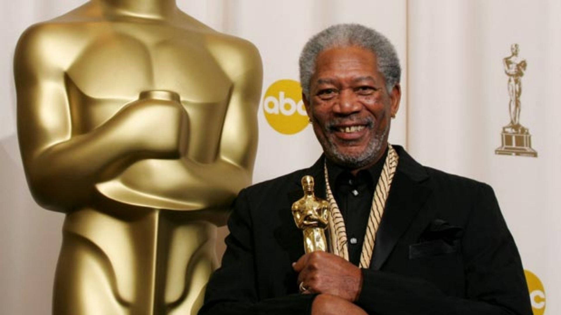 Morgan Freeman 桌布 壁紙 — 摩根費里曼在正式頒獎典禮上自豪地微笑，手持奧斯卡獎座，旁邊有一個巨大的金色奧斯卡複製品。