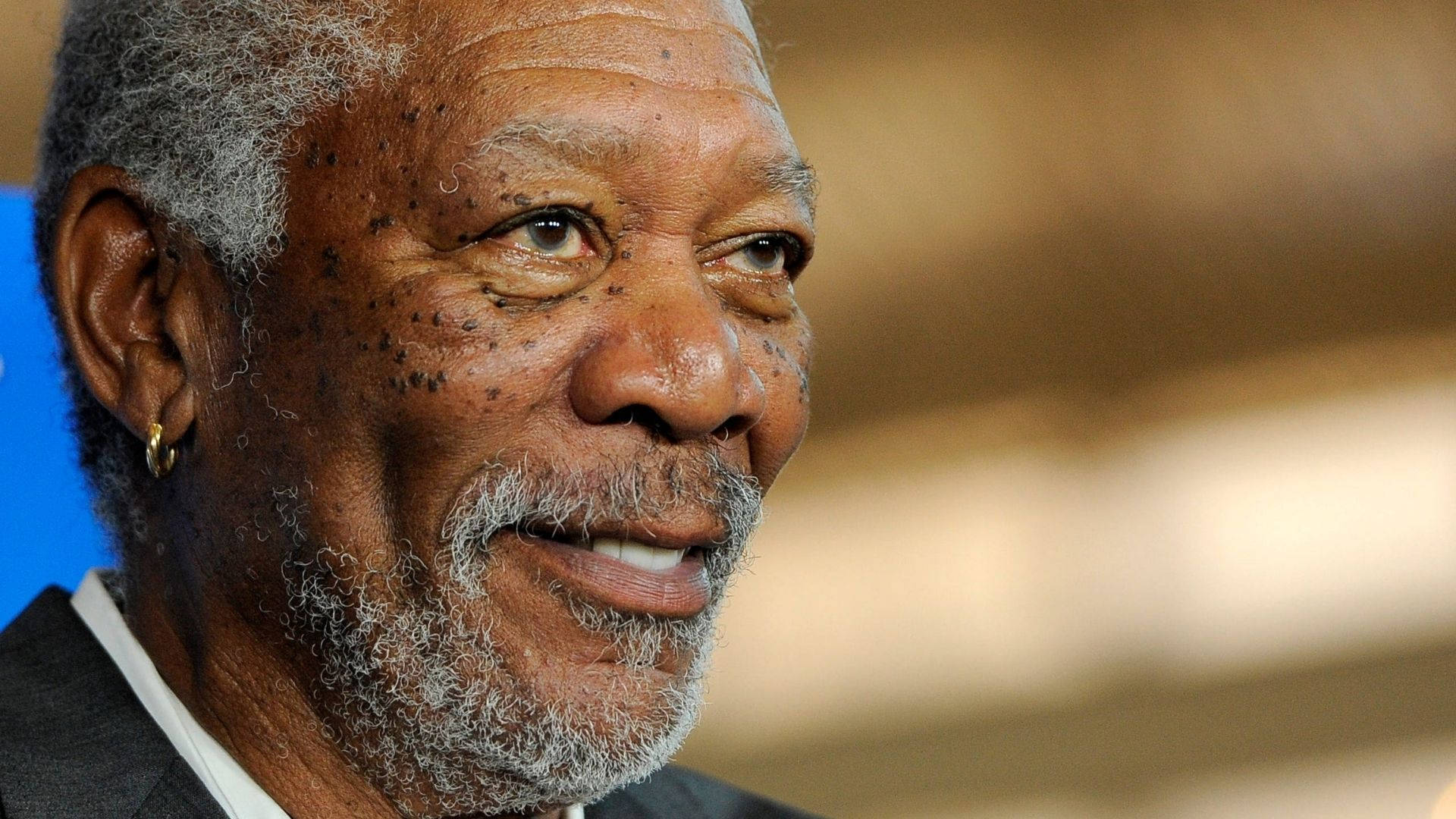 Morgan Freeman 桌布 壁紙 — 摩根費里曼的特寫肖像，他留著灰色鬍鬚並戴著金耳環，在模糊背景前溫暖微笑。