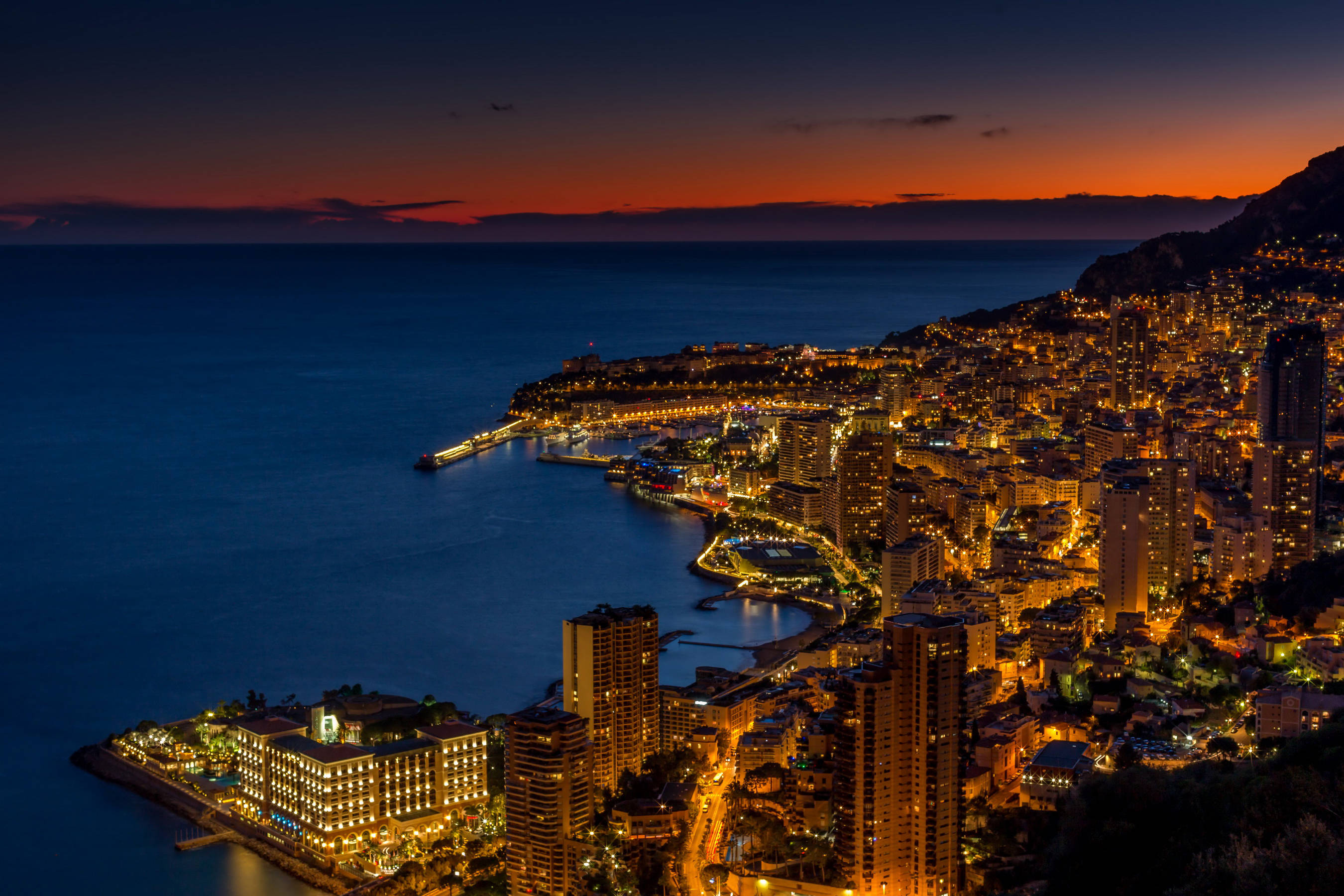 Monaco 桌布 壁紙 — 摩納哥海岸城市景觀在暮色中閃耀著金色燈光，映襯著深藍色的海洋和火紅色的天空，景色令人驚嘆。