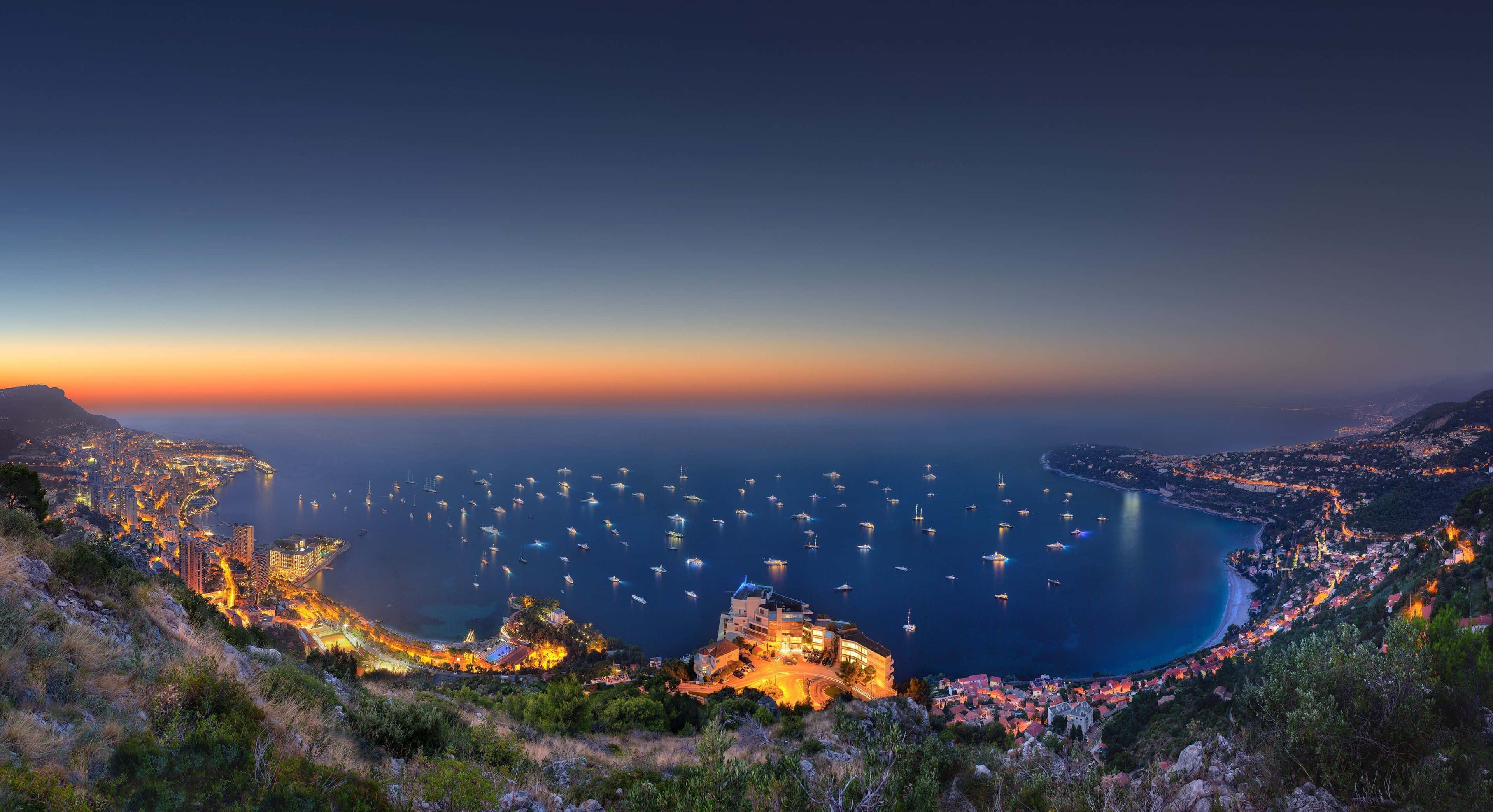Europe 桌布 壁紙 — 摩納哥海岸線的黃昏全景，城市燈光閃爍，深藍色海灣中停泊著眾多帆船。