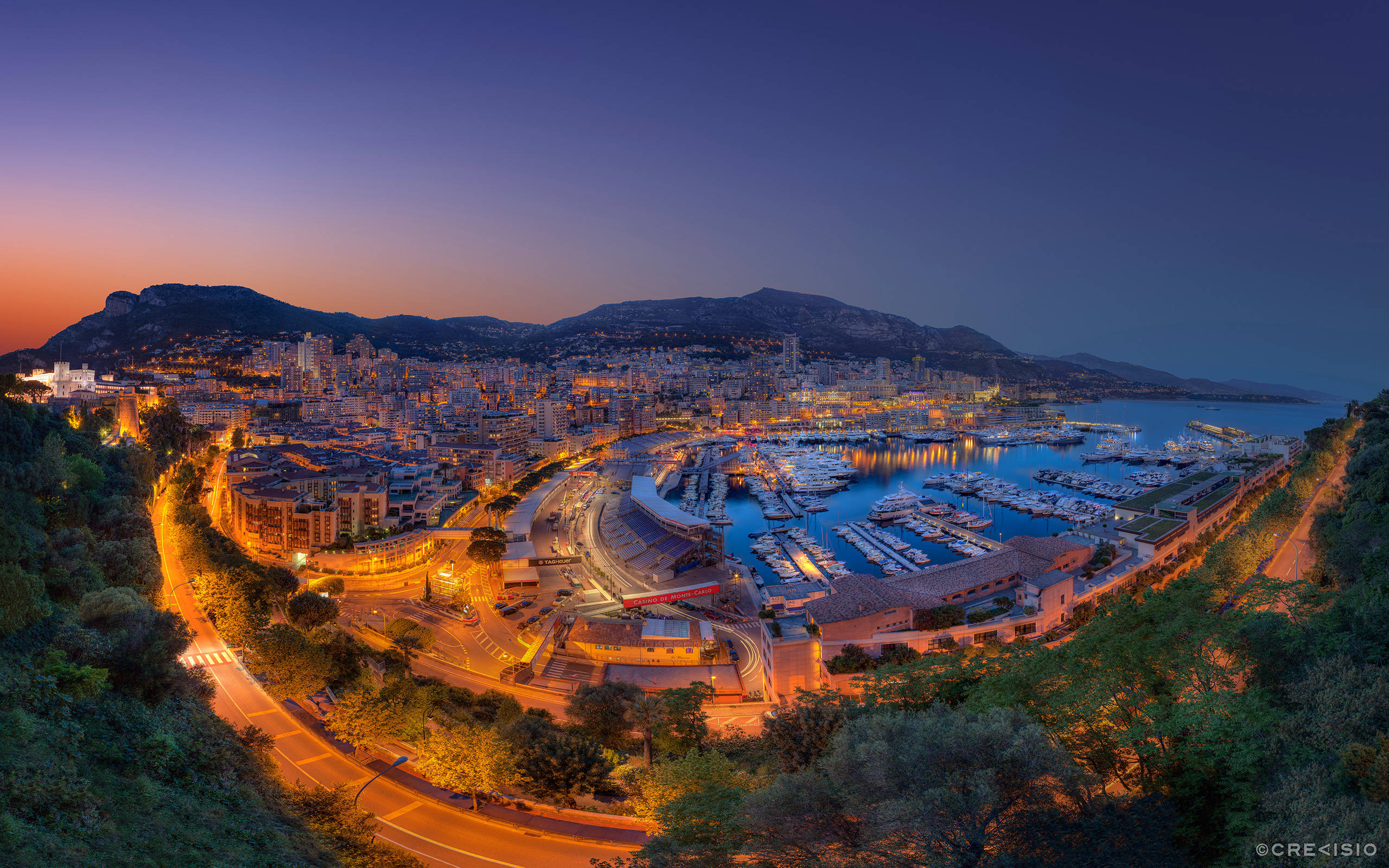 Monaco 桌布 壁紙 — 摩納哥港口和城市景觀的全景黃昏景色，溫暖的燈光在深紫色天空下閃耀。