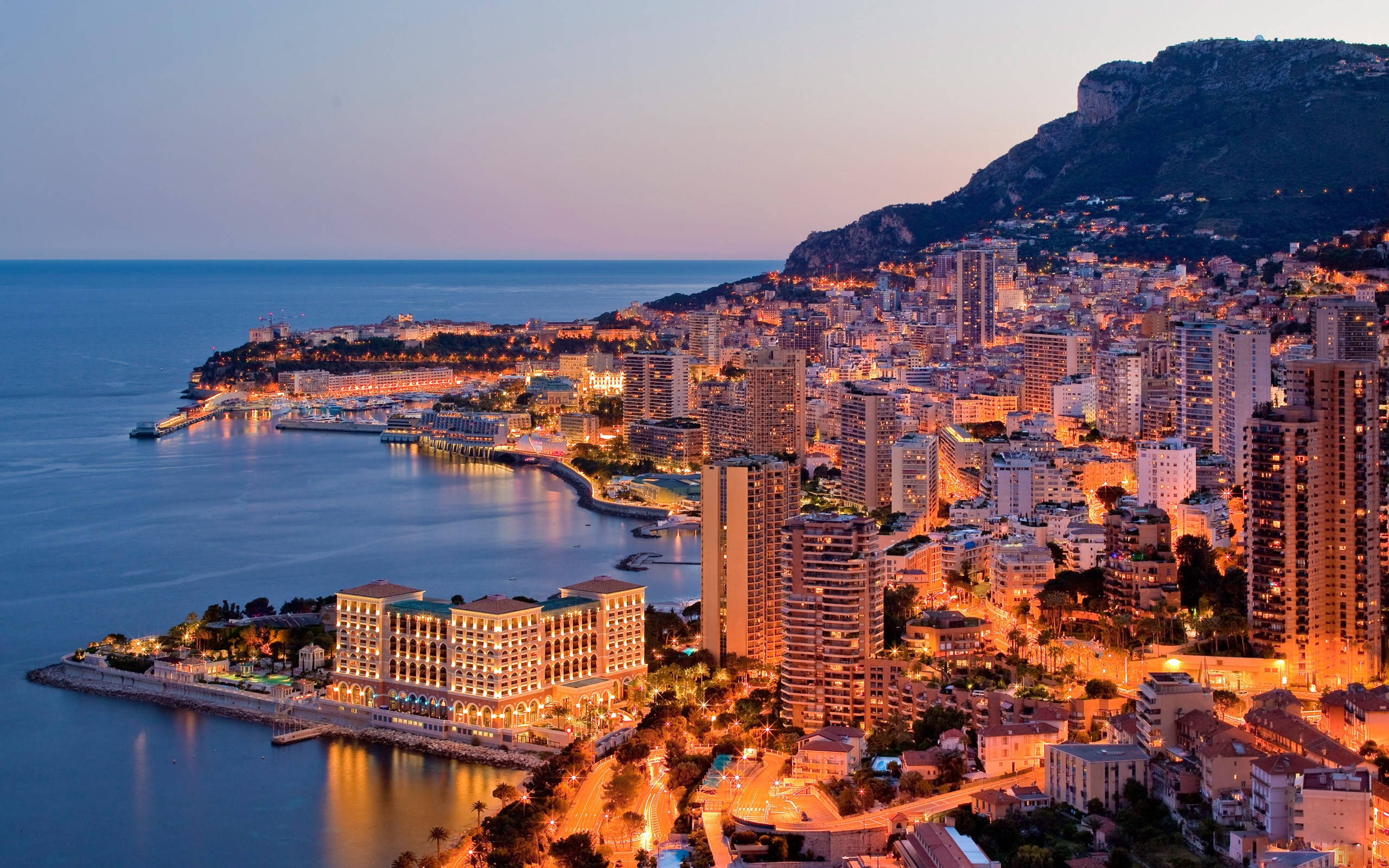 Monaco 桌布 壁紙 — 摩納哥黃昏時的鳥瞰圖，閃爍的城市燈光映照在平靜的海面和岩石山坡上。