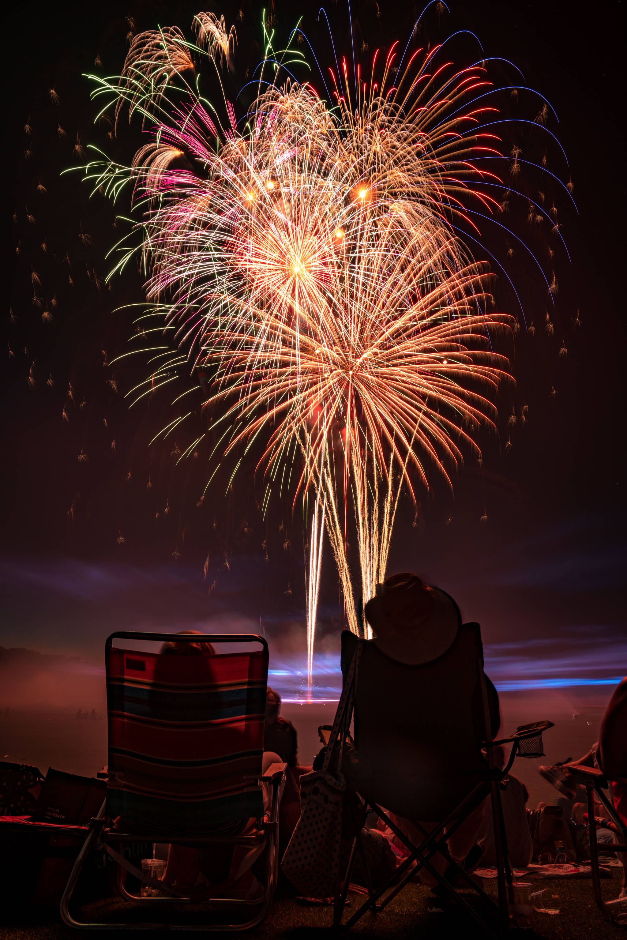 Fireworks 桌布 壁紙 — 摺疊椅上的剪影觀眾觀看紅色、綠色和金色煙火在夜空中綻放耀眼的光芒。