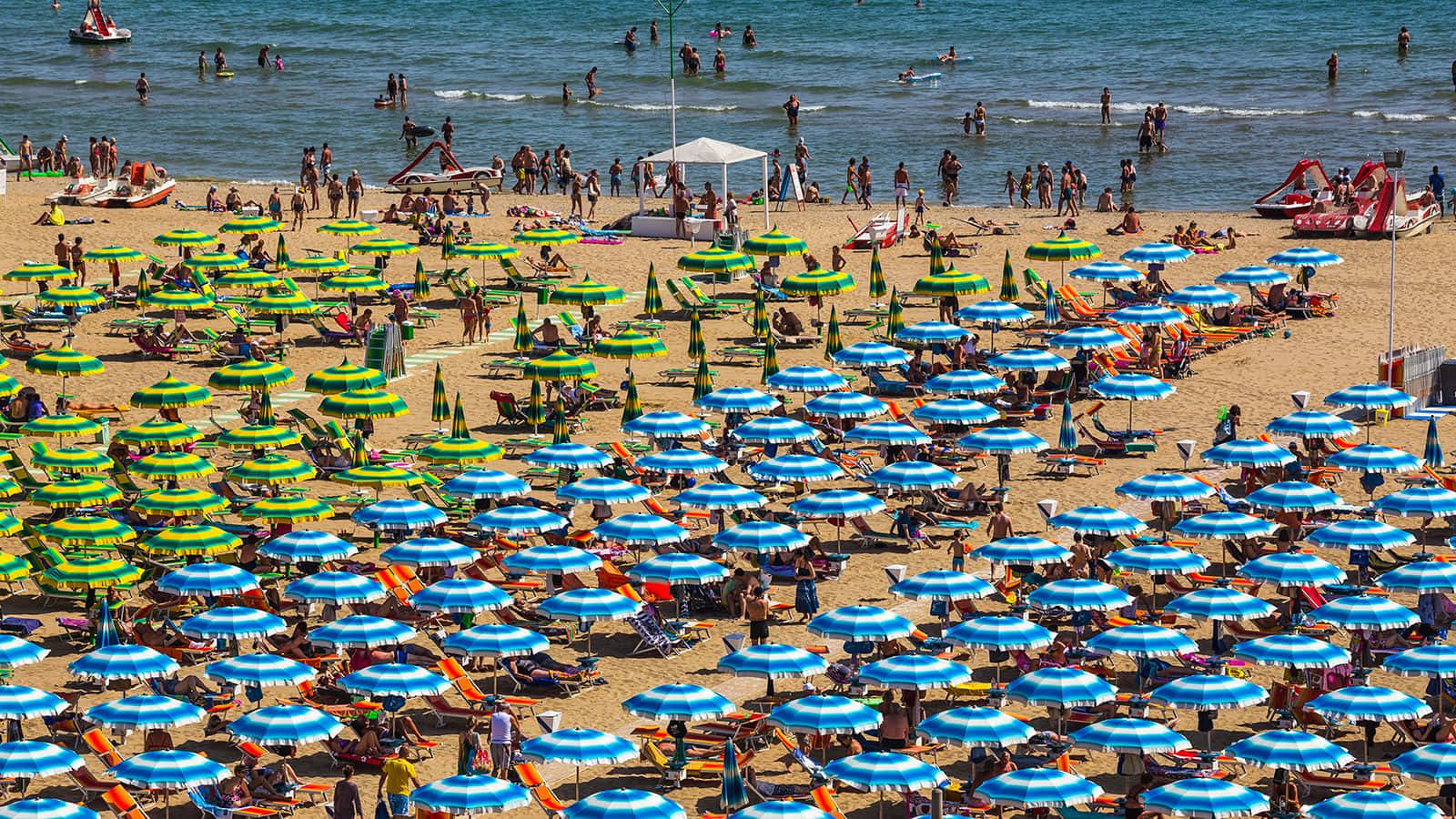 Italian Beach 桌布 壁紙 — 擁擠海灘的鳥瞰圖，藍綠條紋遮陽傘點綴在金色沙灘上，人們在明媚陽光下游泳和曬太陽。