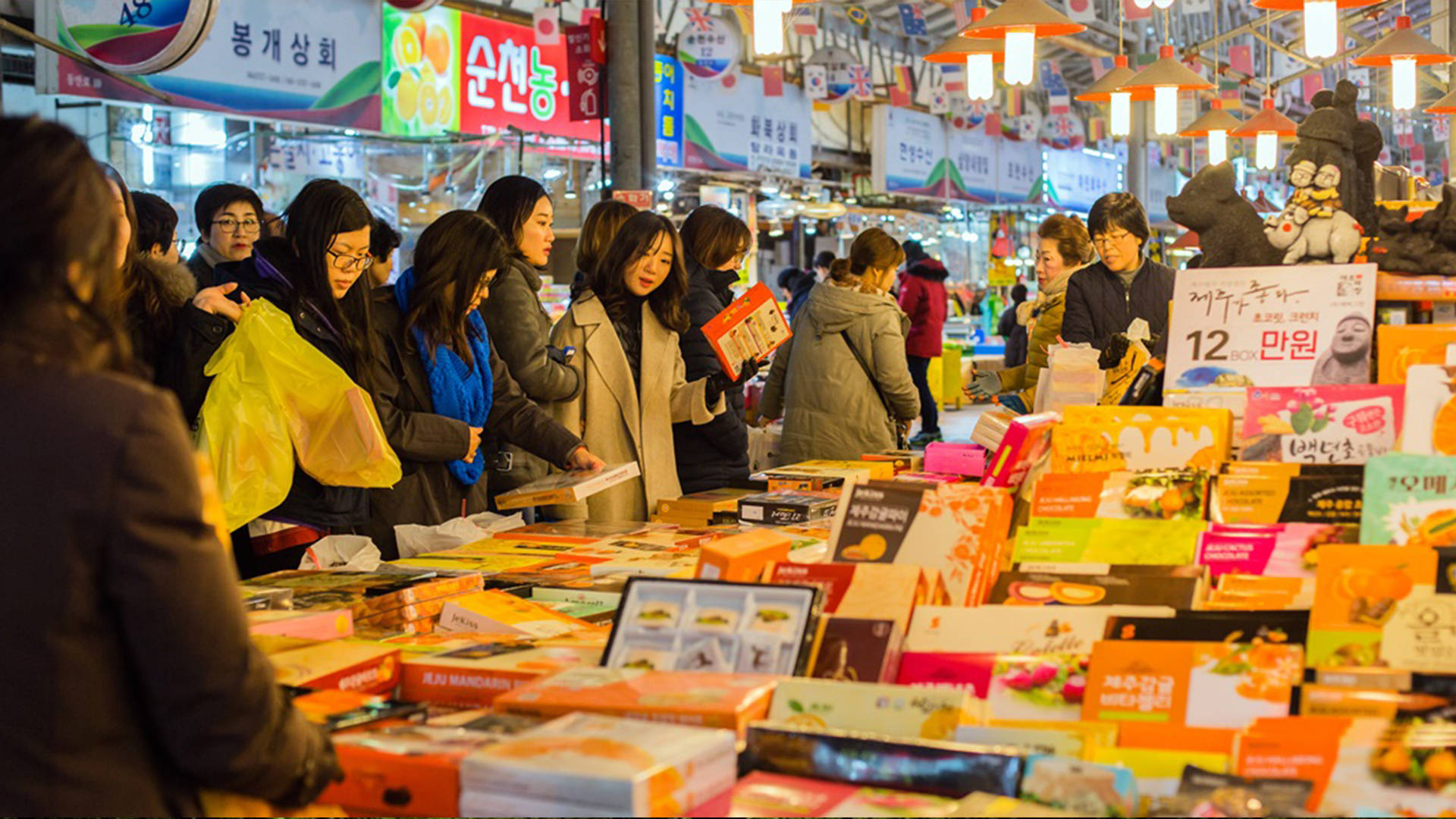 Jeju 桌布 壁紙 — 擁擠的夜市攤位堆滿了色彩繽紛的包裝商品，購物者在明亮的吊燈下瀏覽。