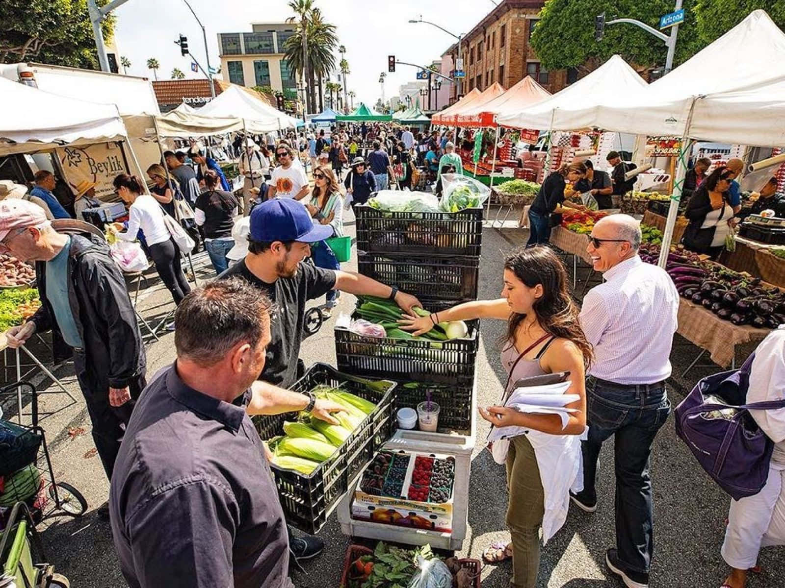 Farmers Market 桌布 壁紙 — 擁擠的戶外農貿市場，人們在陽光明媚的街道上白色帳篷下瀏覽新鮮農產品。