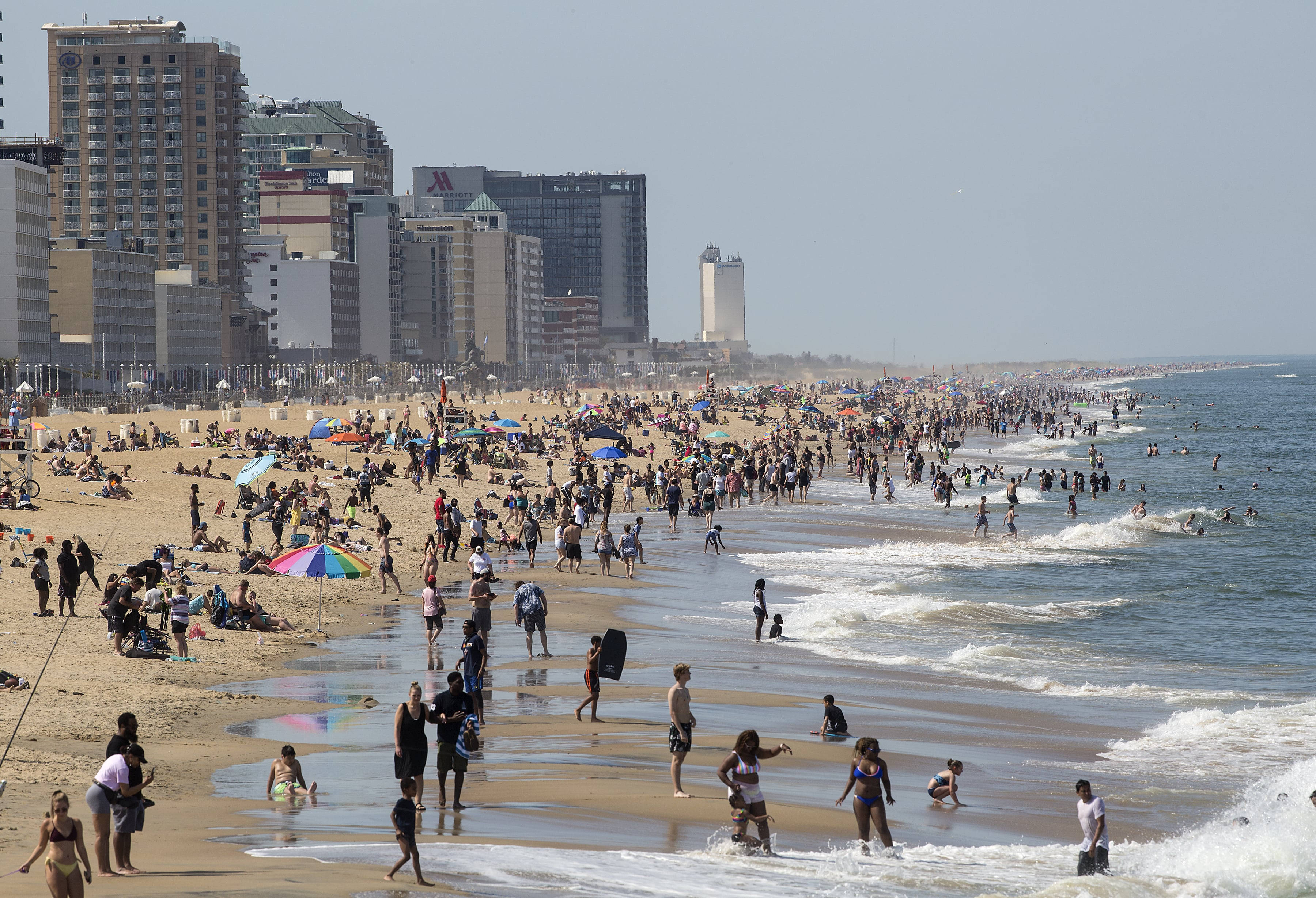 海洋 桌布 壁紙 — 擁擠的海灘，日光浴者和游泳者在明亮的日光下享受海浪，背景是城市天際線。