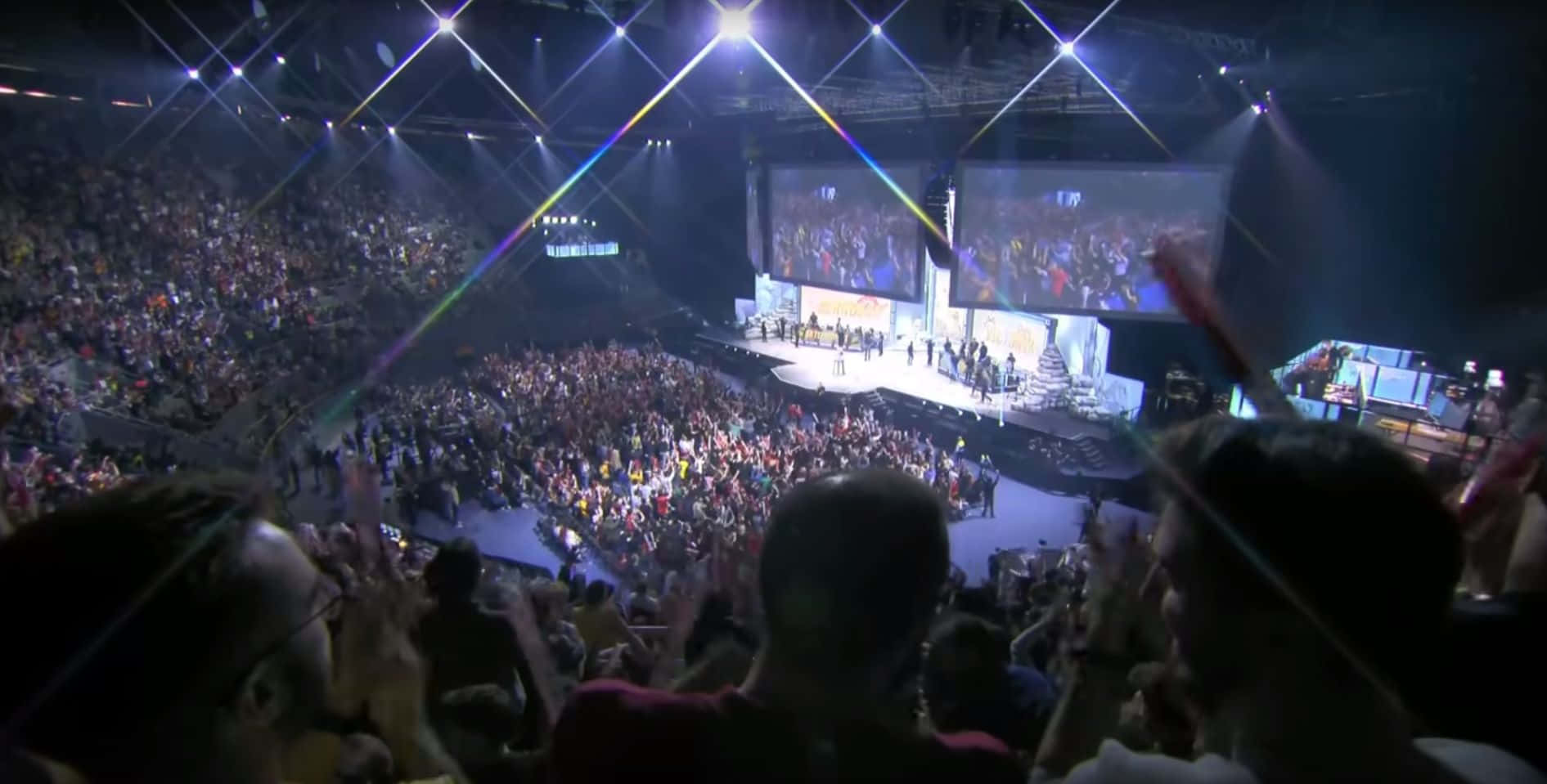 Esports 桌布 壁紙 — 擠滿的場館人群在現場演唱會期間，在帶有彩虹鏡頭眩光的耀眼舞台燈光下歡呼。