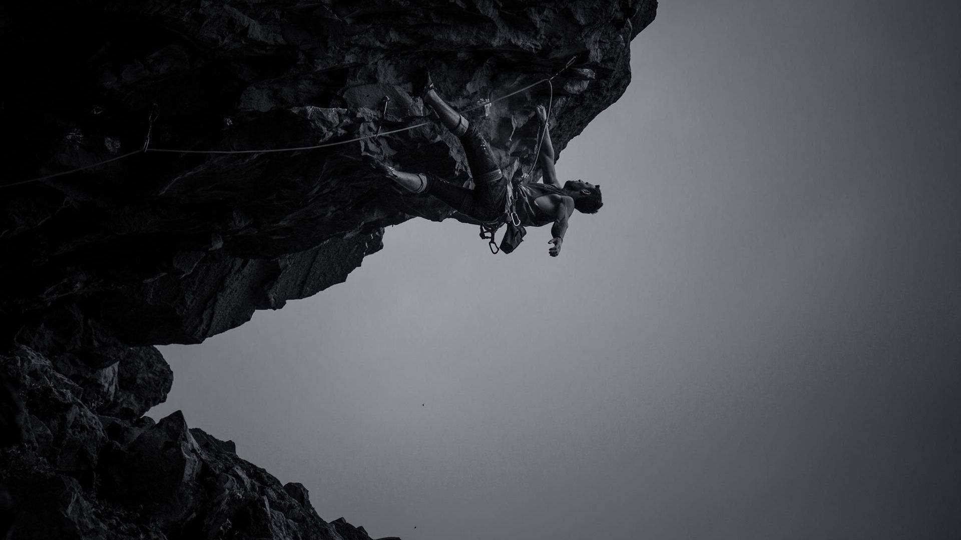 Climbing 桌布 壁紙 — 攀岩者懸掛在岩石突出物下方，背景是柔和天空，以戲劇性的低角度黑白照片拍攝。