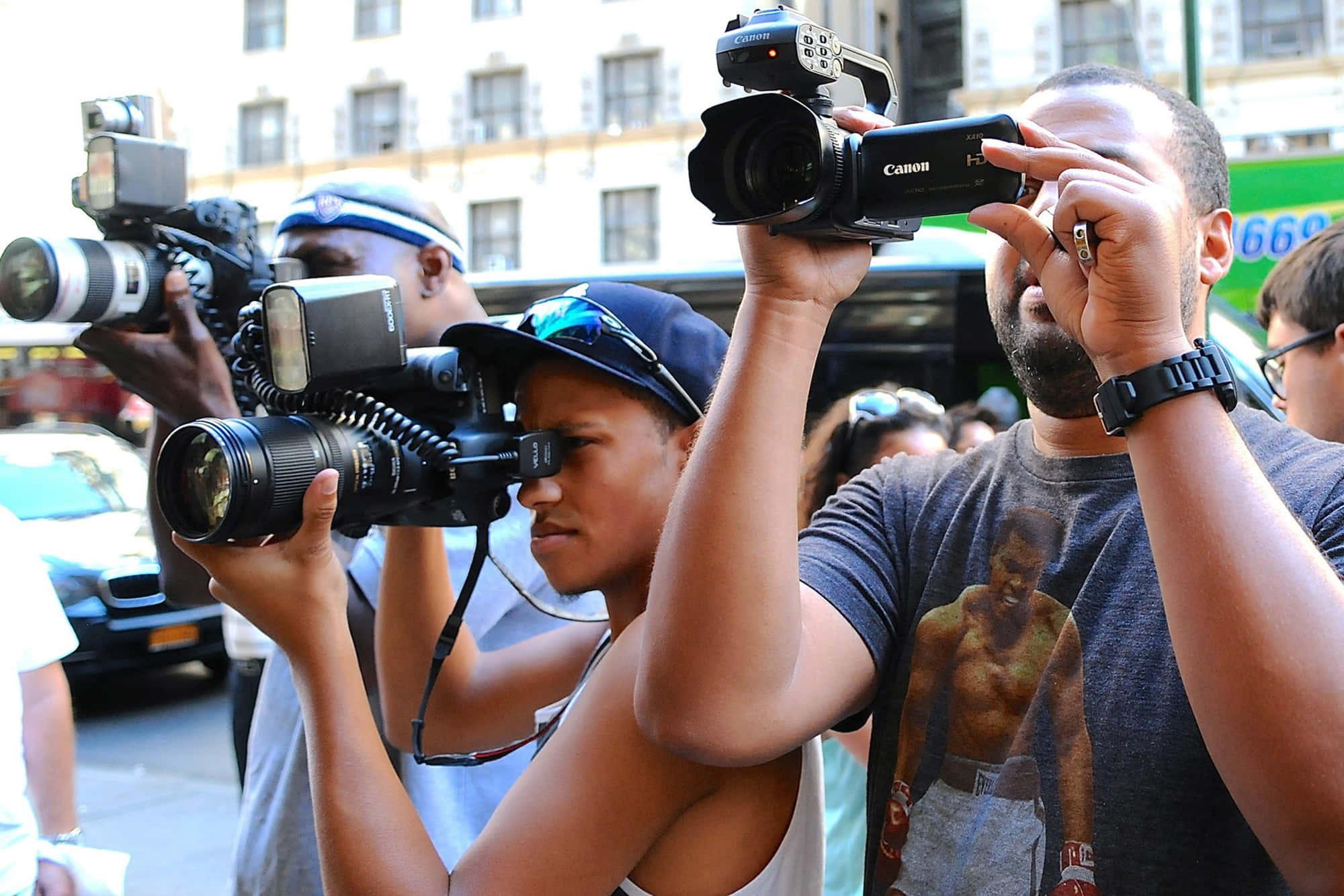 Paparazzi 桌布 壁紙 — 攝影師們帶著大型相機，在繁忙的城市街道上捕捉瞬間，背景有建築物和汽車。