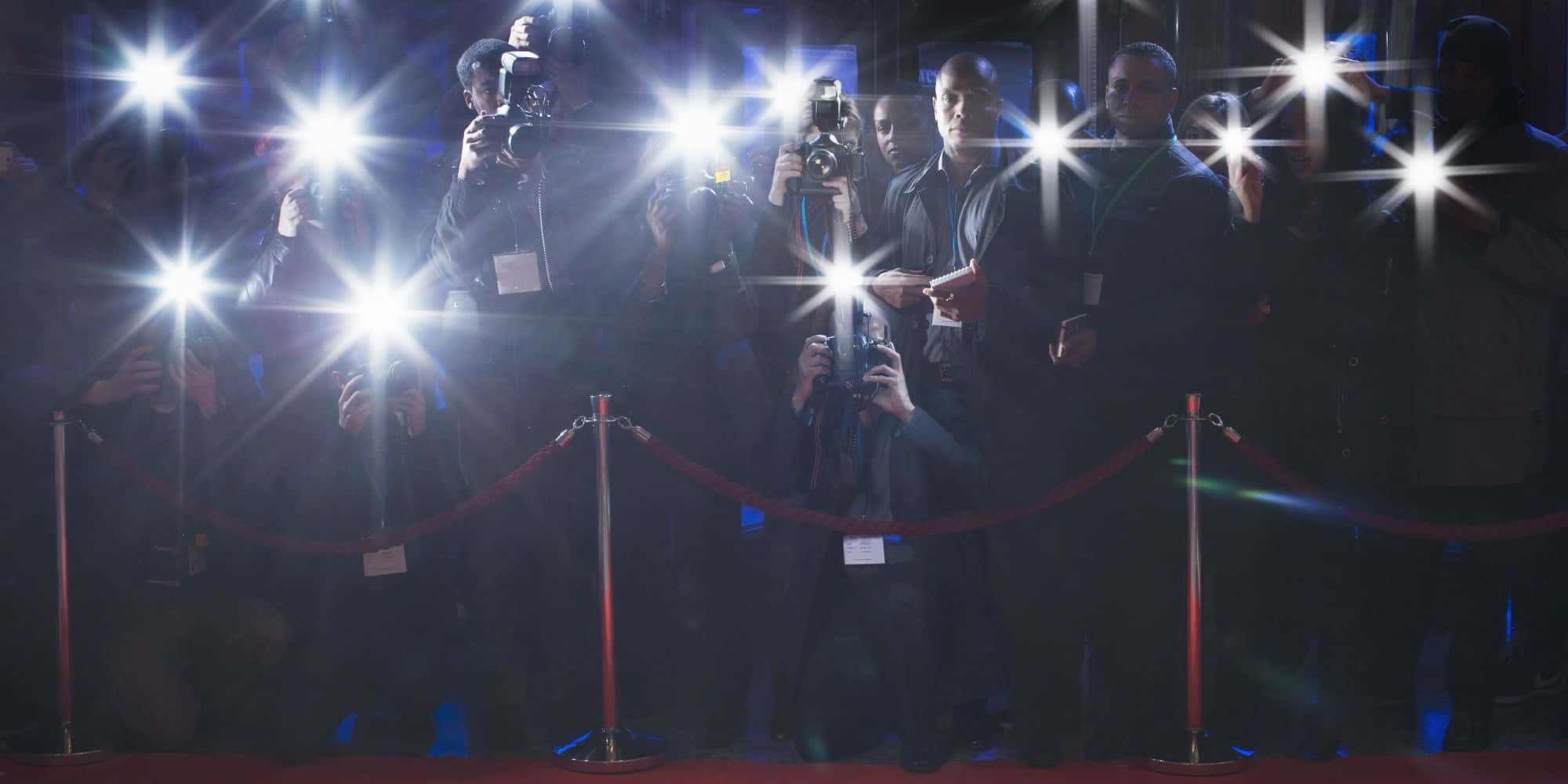 Paparazzi 桌布 壁紙 — 攝影師們帶著閃光燈的相機，在明亮的攝影棚燈光下，擠在天鵝絨繩索後面的紅地毯上。