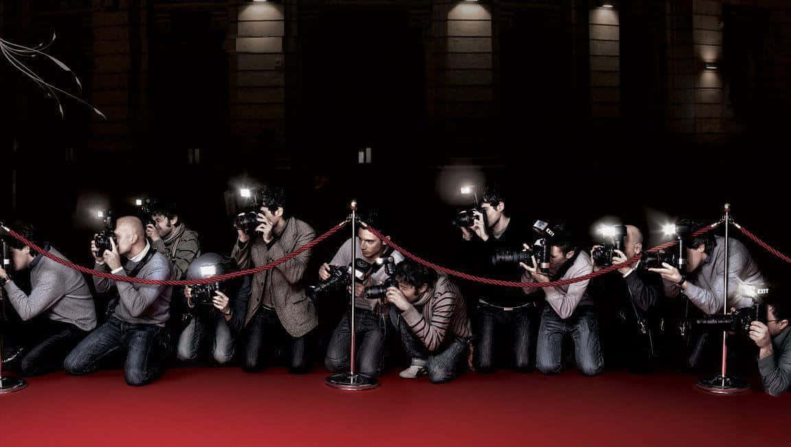 Paparazzi 桌布 壁紙 — 攝影師跪在天鵝絨繩索後面的紅地毯上，在光線昏暗的場地中將閃光燈相機對準一個看不見的主體。