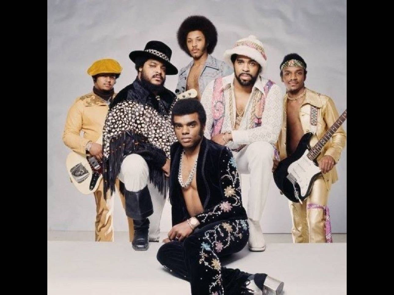 音樂 桌布 壁紙 — 放克樂團 Earth, Wind & Fire 的復古工作室肖像，穿著華麗的 1970 年代服裝，在中性背景前拿著吉他擺姿勢。