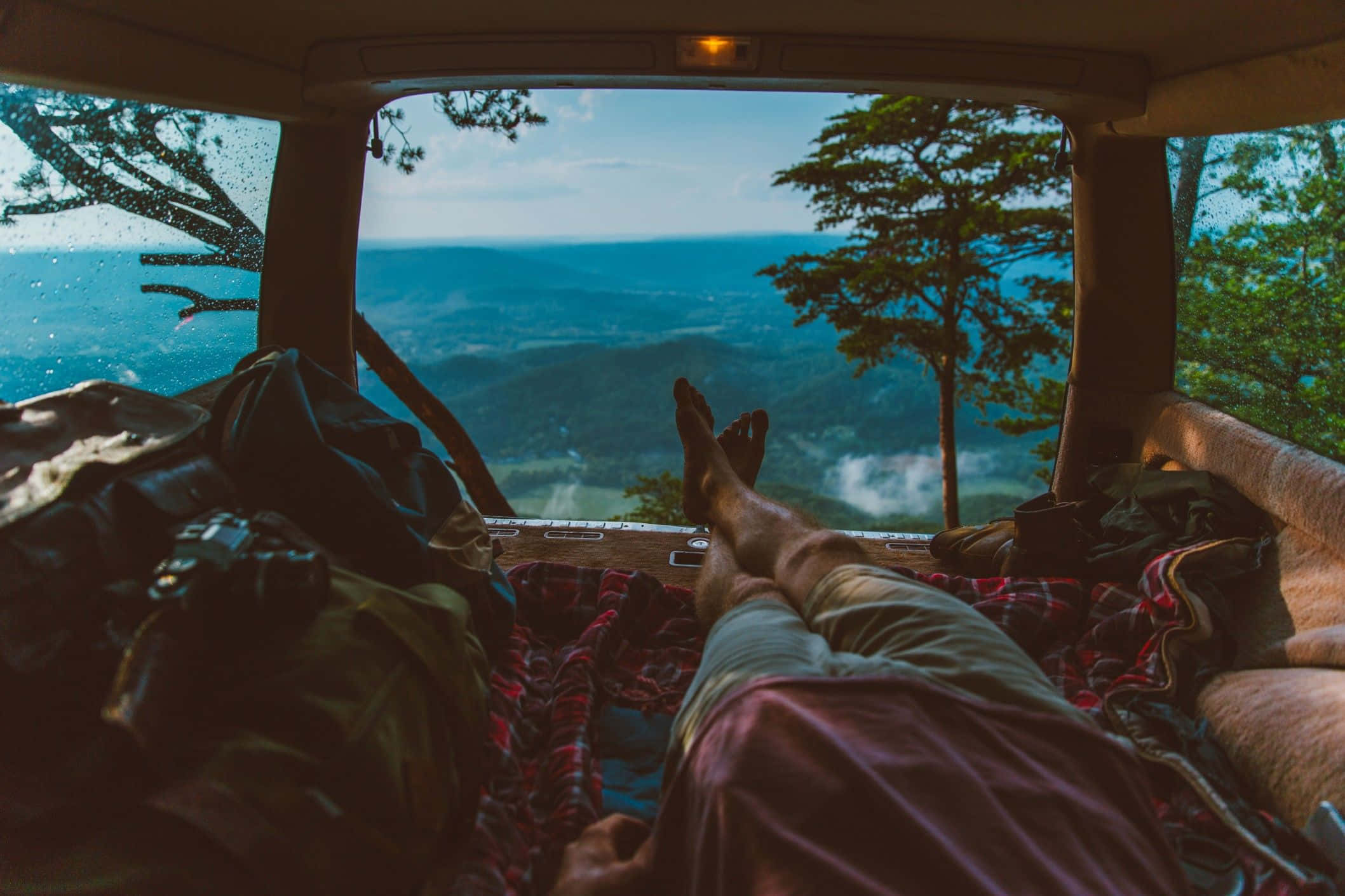 Minimalist Camping 桌布 壁紙 — 放鬆的旅客赤腳躺在露營車床上，透過打開的後窗凝視著霧氣瀰漫的山谷。