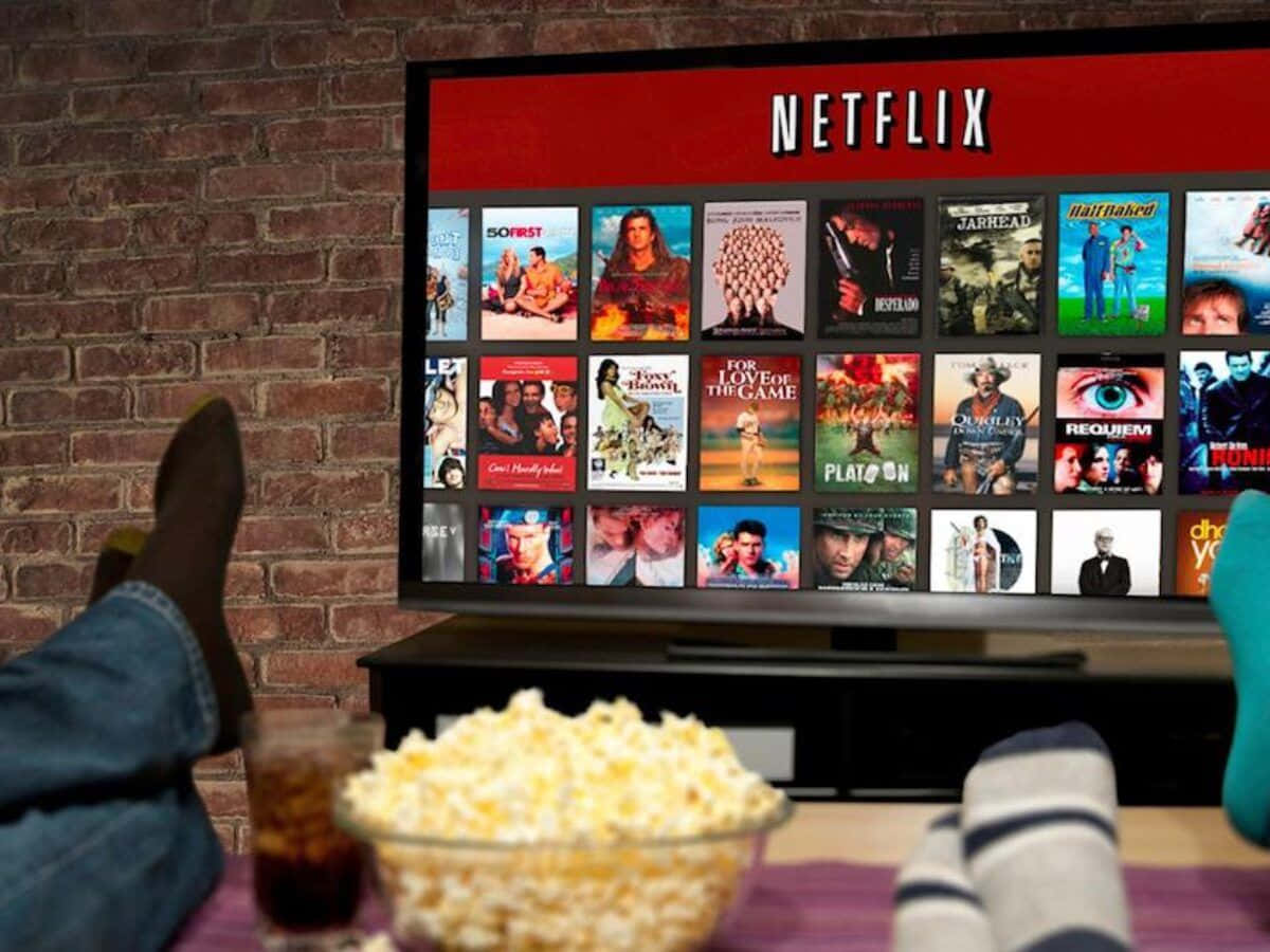 Netflix 桌布 壁紙 — 放鬆的雙腳穿著牛仔褲擱在桌上，旁邊有爆米花和汽水，電視在磚牆背景下顯示Netflix首頁。