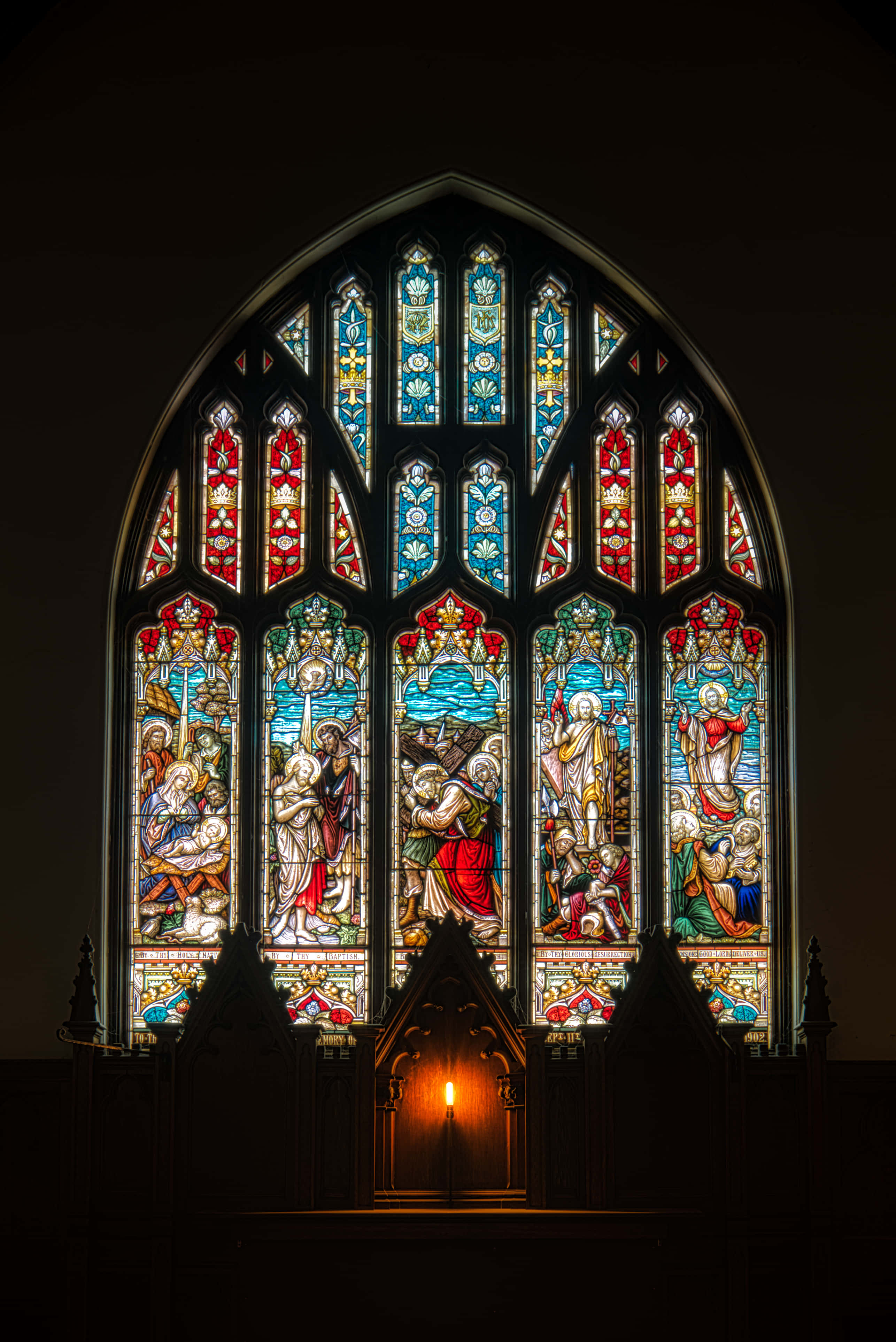 Stained Glass 桌布 壁紙 — 教堂內一幅巨大的彩繪玻璃窗，在深色石牆上投射出色彩繽紛的聖經場景，下方有一根蠟燭在發光。