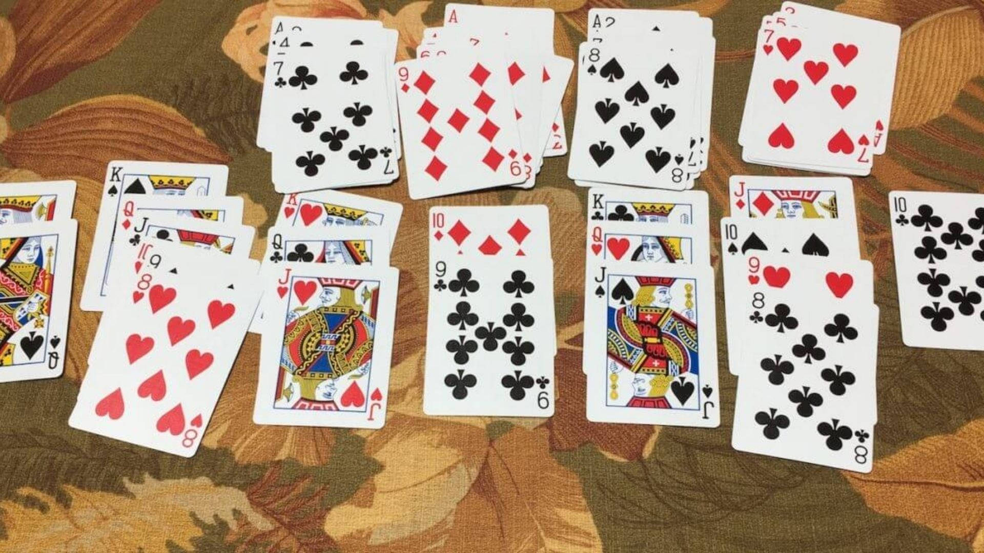 Solitaire 桌布 壁紙 — 散落的紅色和黑色花色撲克牌放在有圖案的棕色和米色布料表面上。