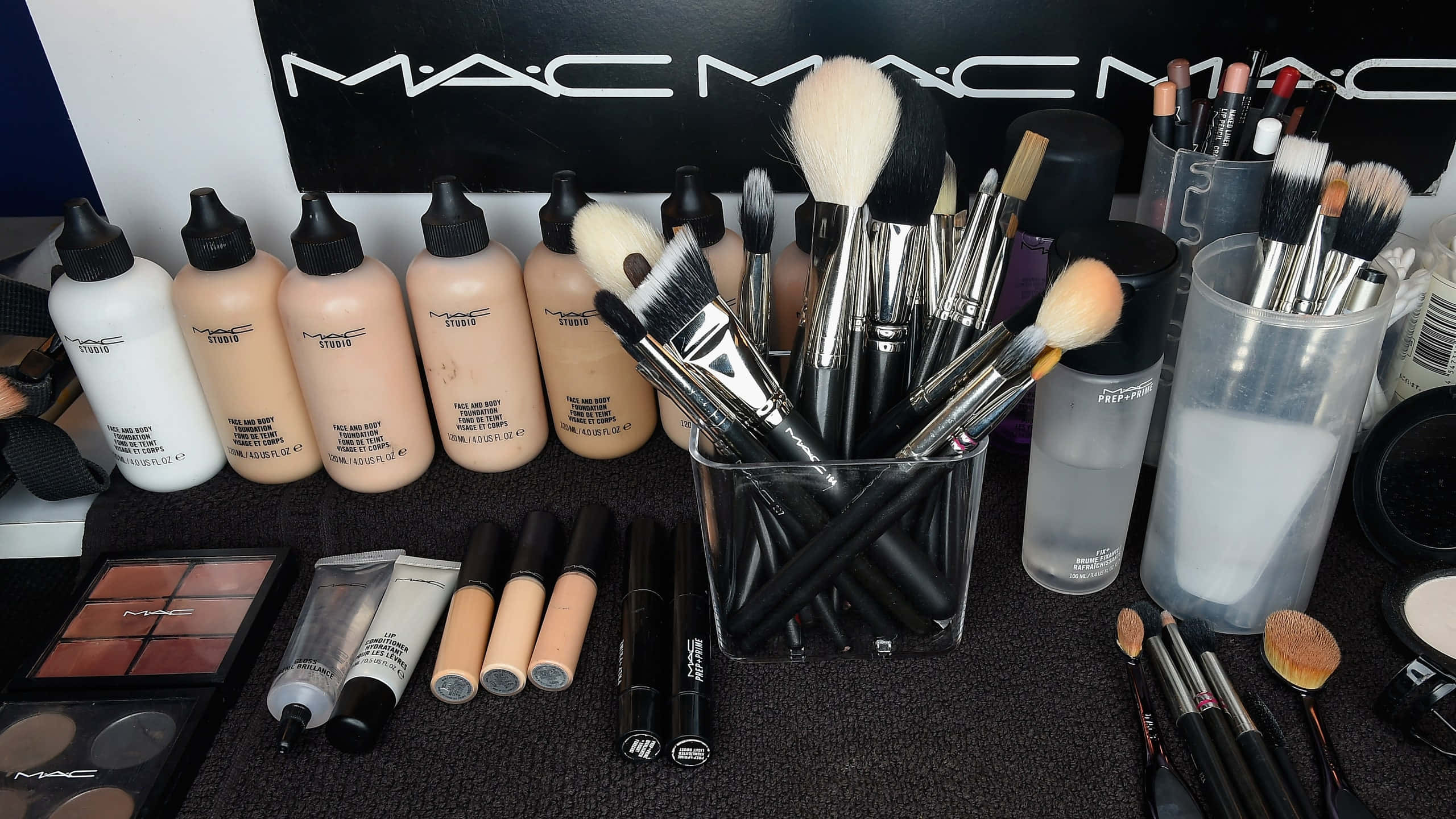 Mac Cosmetics 桌布 壁紙 — 整齊排列的MAC化妝品系列，包括粉底瓶、刷子和眼影盤，放在深色紋理表面上。