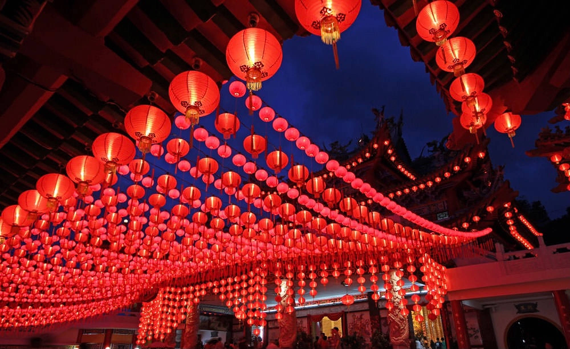 Kuala 桌布 壁紙 — 數千盞發光的紅色燈籠如瀑布般懸掛在傳統寺廟屋頂上，映襯著深藍色的暮色天空。