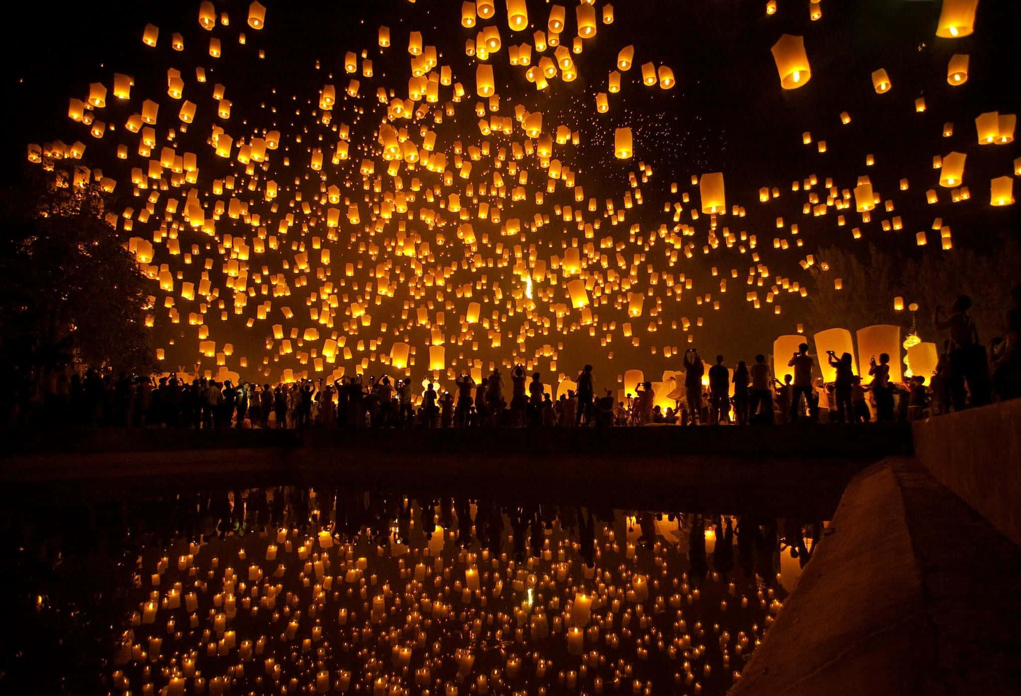 Lantern 桌布 壁紙 — 數百盞發光的天燈從人群中升起，人群在黑暗夜空的映襯下形成剪影，夜空倒映在下方水中。