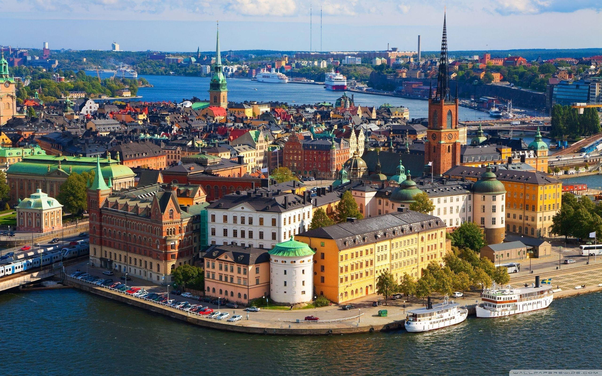 Sweden 桌布 壁紙 — 斯德哥爾摩歷史區的鳥瞰圖，色彩繽紛的建築物、尖塔和船隻在藍色水面上，天空晴朗。