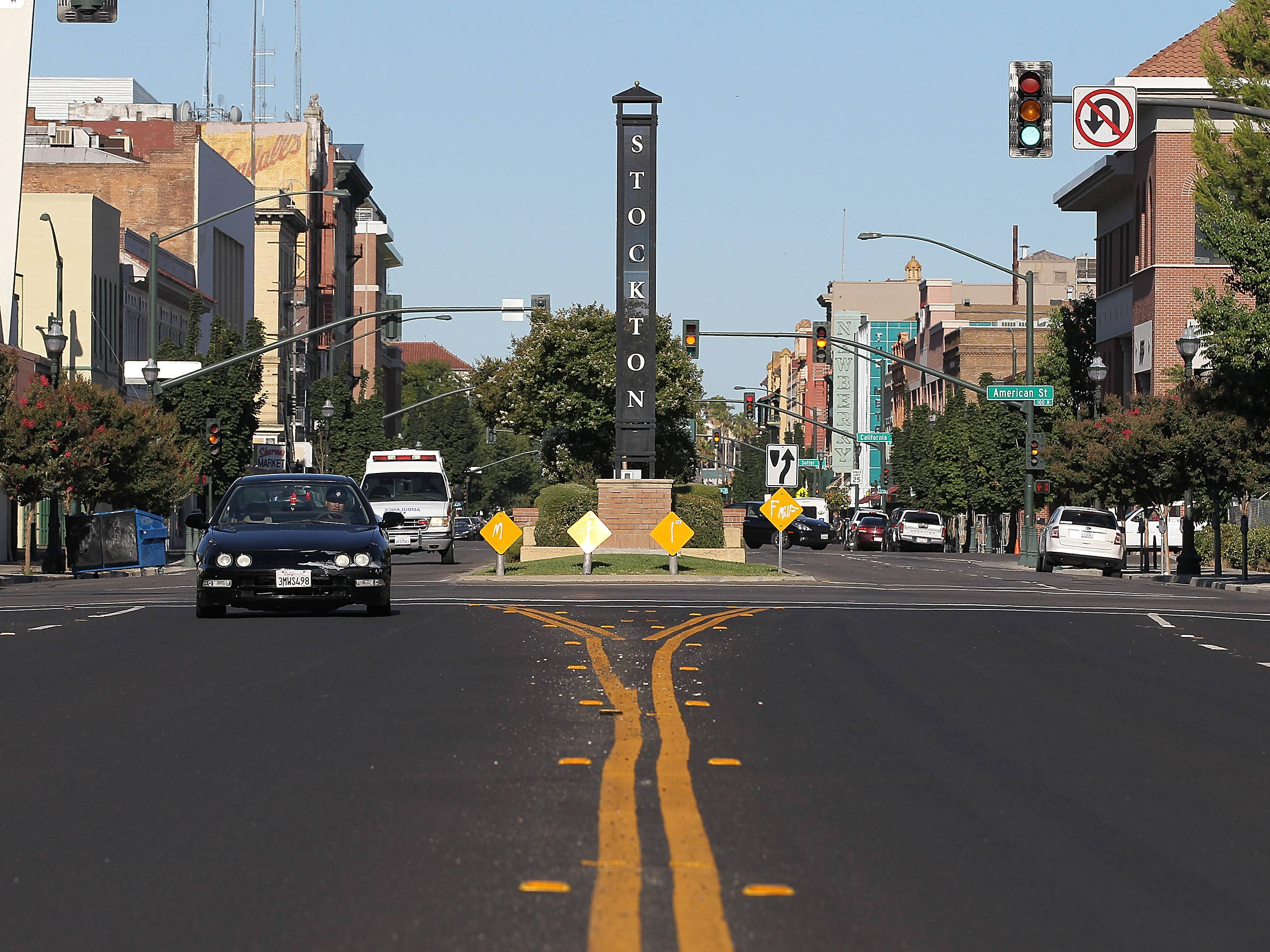 City Street 桌布 壁紙 — 斯托克頓陽光明媚的城市街景，汽車在交通號誌下行駛，一個高大的黑色標誌標示著城市入口。