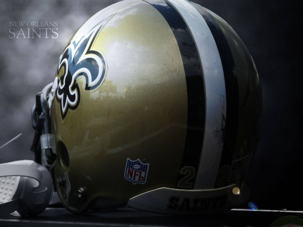 聖徒 桌布 壁紙 — 新奧爾良聖徒隊NFL美式足球頭盔特寫，帶有金色、黑色和白色條紋以及百合花徽標，背景為煙霧瀰漫的黑暗。