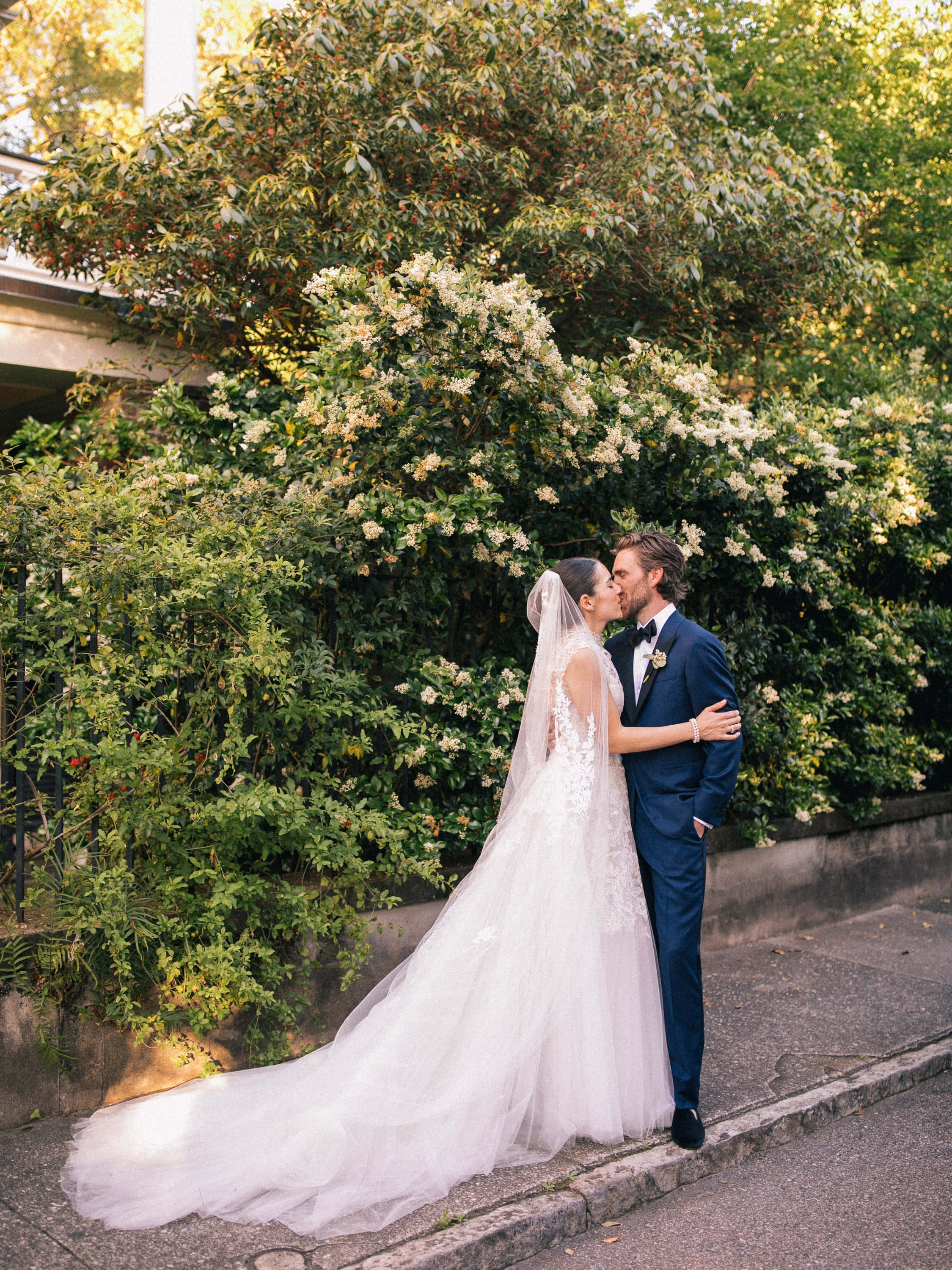 Garden Wedding 桌布 壁紙 — 新娘和新郎在人行道上溫柔親吻，旁邊是開滿白花的茂密綠色樹籬。