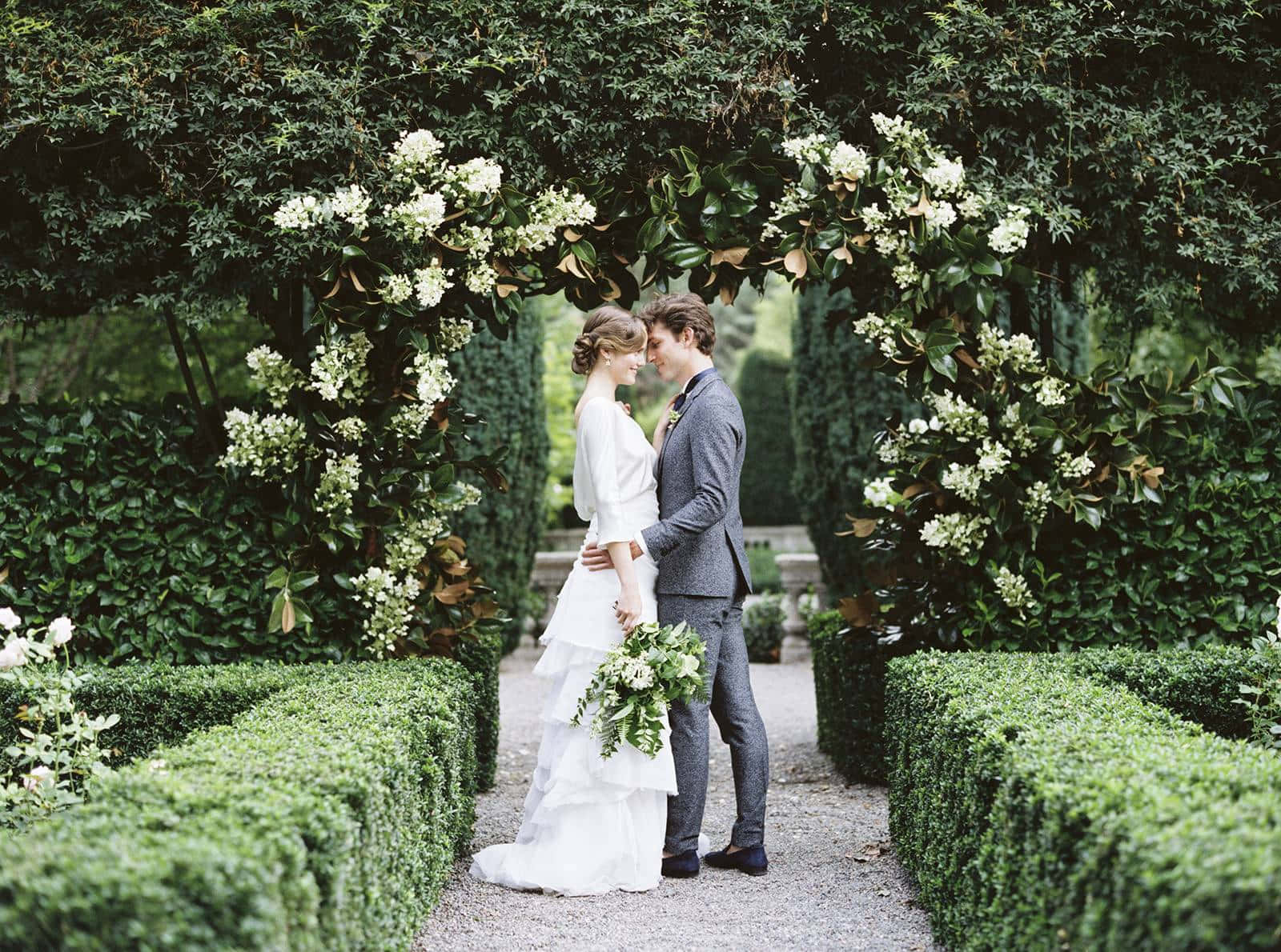 Garden Wedding 桌布 壁紙 — 新娘和新郎在修剪整齊的花園中，於一個佈滿白花的茂盛綠色拱門下擁抱。