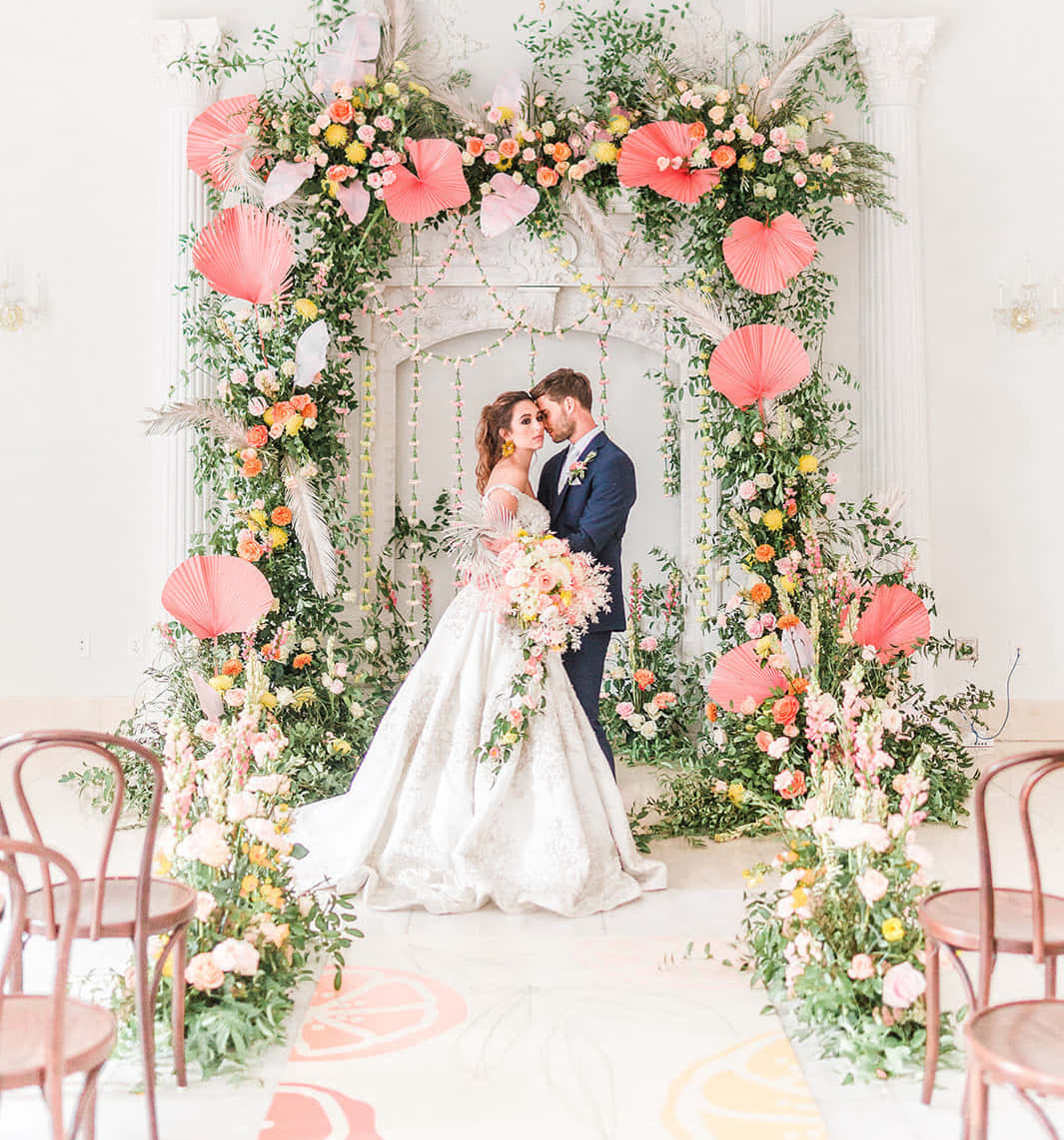 Garden Wedding 桌布 壁紙 — 新娘和新郎在明亮的婚禮場地中，在一個裝飾著粉紅色紙扇和綠植的茂盛花拱門下擁抱。