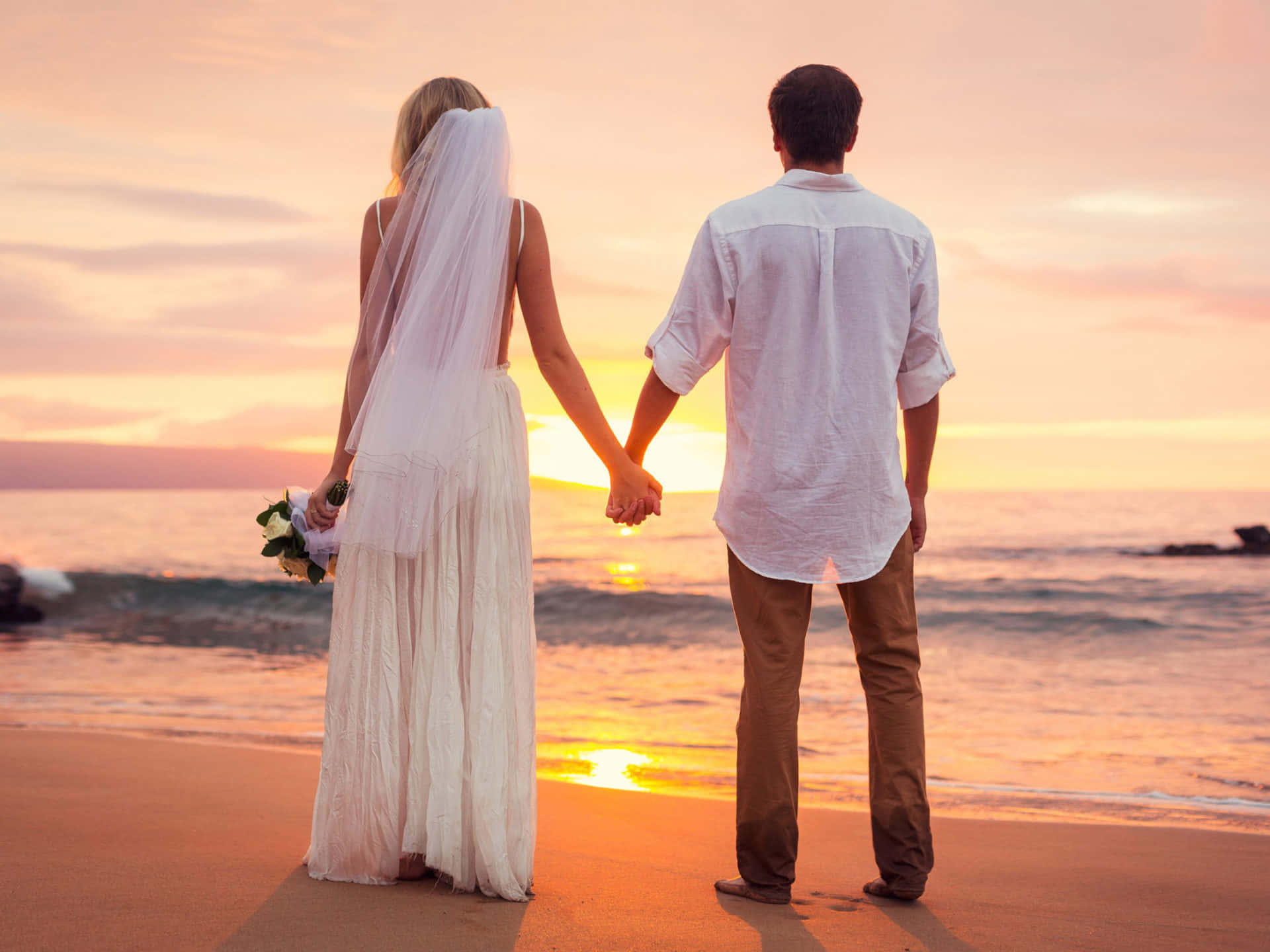 Beach Lovers 桌布 壁紙 — 新娘和新郎在海灘上牽手，夕陽在他們身後落下，將金色的光芒灑在海面上。