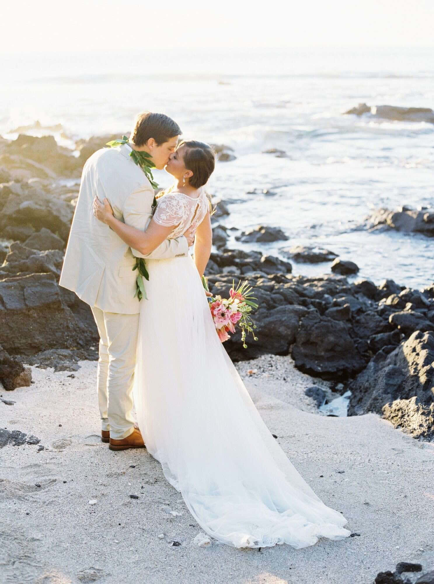 Beach Wedding 桌布 壁紙 — 新娘和新郎在陽光明媚的岩石海灘上溫柔親吻，身後海浪拍打。