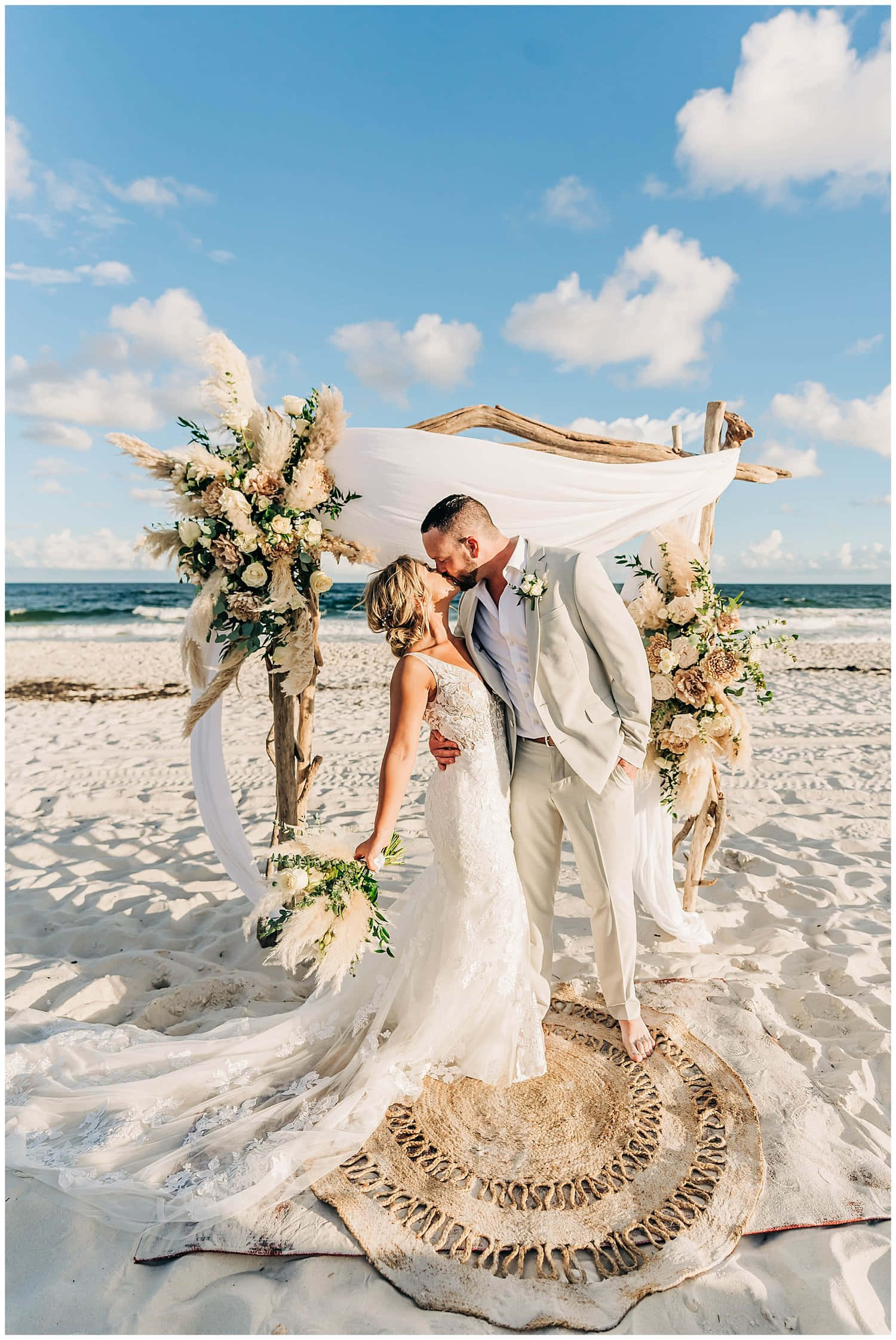 Beach Wedding 桌布 壁紙 — 新娘和新郎在陽光明媚的海灘上，在佈滿鮮花的木拱門下接吻，背景是海浪和藍天。