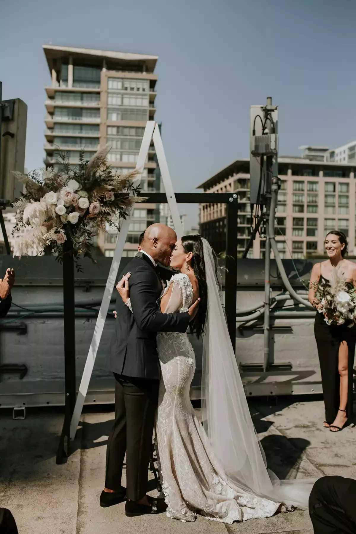 Black And White Wedding 桌布 壁紙 — 新娘和新郎在陽光普照的屋頂婚禮上，在花拱門下接吻，背景是城市建築。