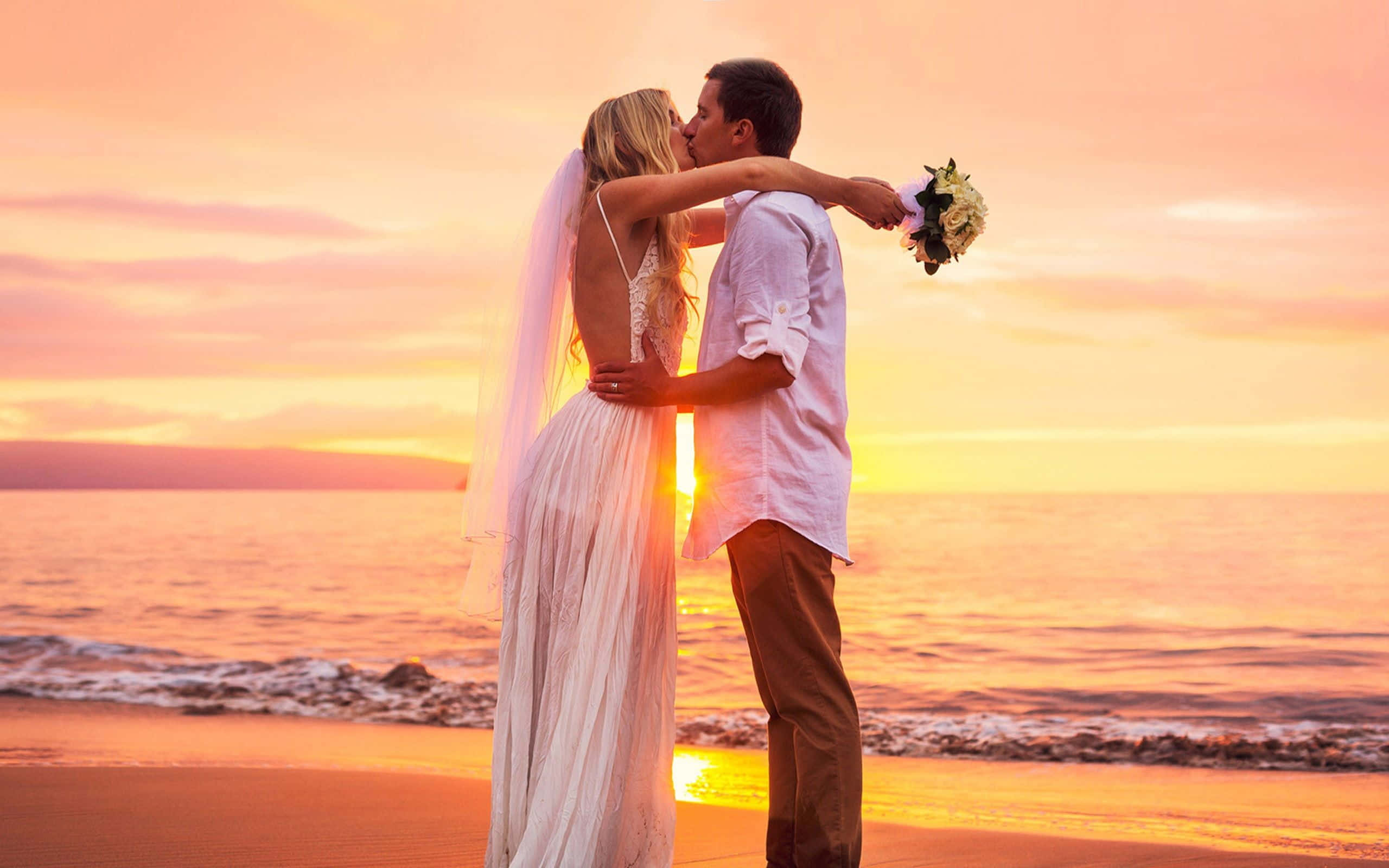 Wedding 桌布 壁紙 — 新娘和新郎在陽光普照的海灘上熱情接吻，金色夕陽的光芒沐浴著海洋和天空。