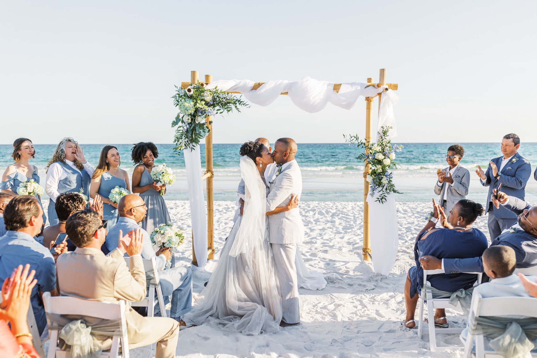 Beach Wedding 桌布 壁紙 — 新娘和新郎在陽光海灘上，於一個裝飾著白色布幔和綠植的木拱門下接吻，賓客們鼓掌。