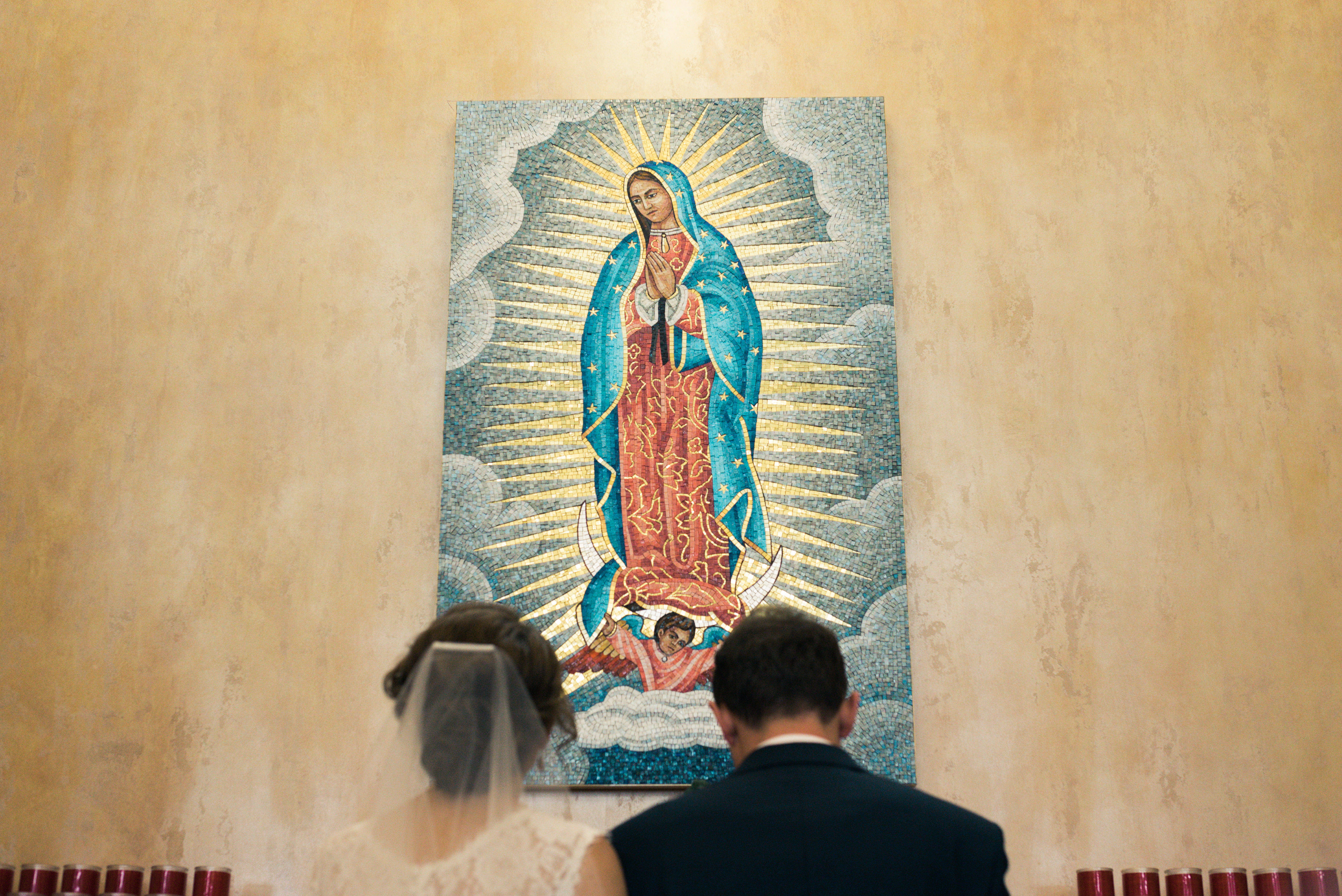 Catholic 桌布 壁紙 — 新娘和新郎站在一幅色彩鮮豔的瓜達盧佩聖母馬賽克前，馬賽克在紋理米色牆壁上散發著金色光芒。