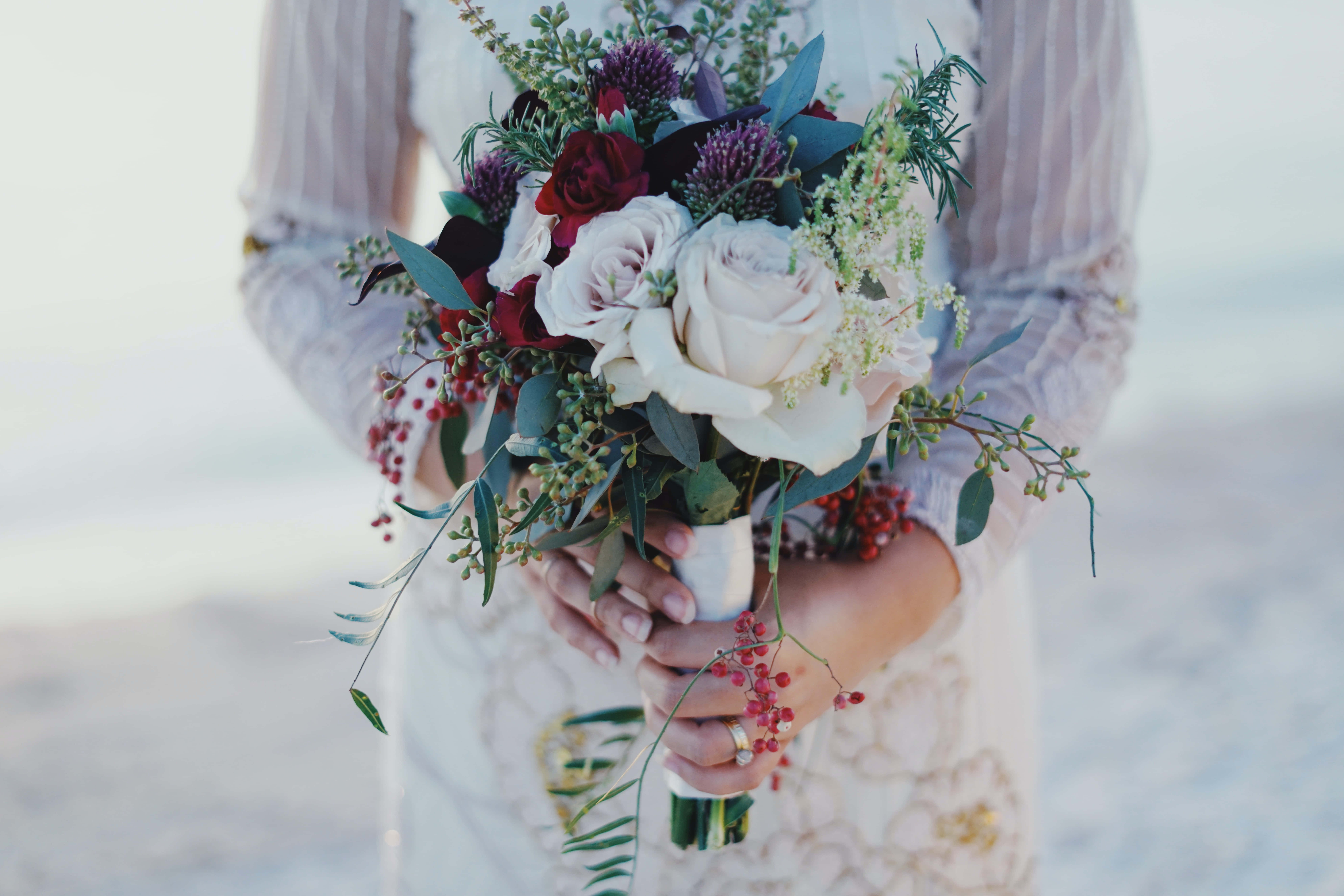 Wedding Bouquet 桌布 壁紙 — 新娘手持一束由白玫瑰、深紅花朵和綠葉組成的豐滿花束，背景是柔和的海灘。