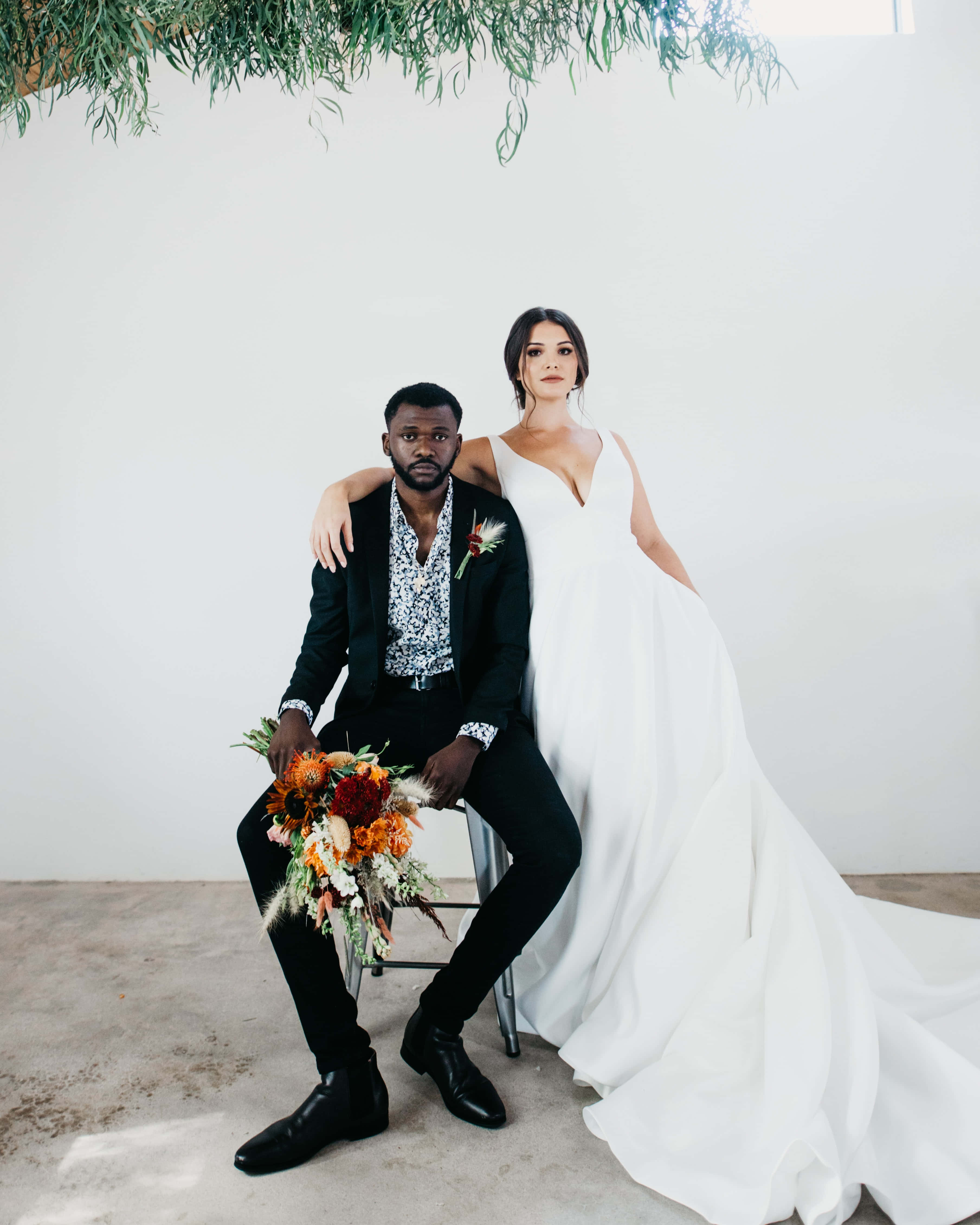 Interracial 桌布 壁紙 — 新娘穿著飄逸的白色婚紗，新郎穿著黑色西裝，在懸掛的綠色植物下，手持鮮豔的秋季花束合影。