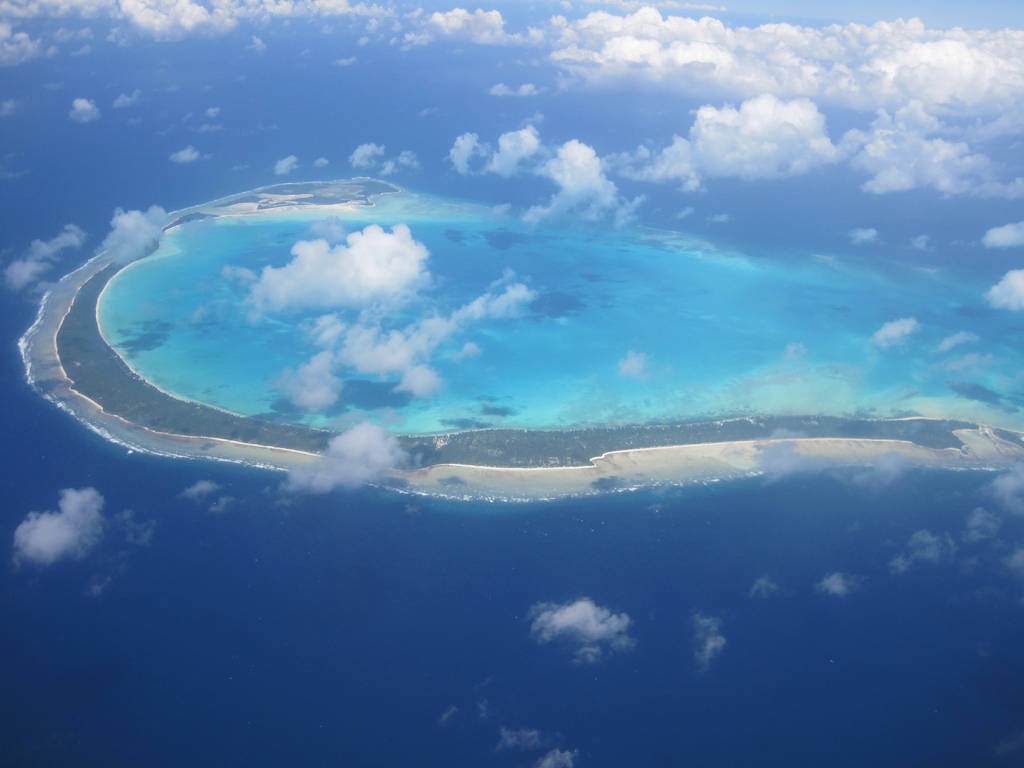 Kiribati 桌布 壁紙 — 新月形熱帶環礁的鳥瞰圖，擁有碧綠色的潟湖和白色沙灘，周圍是深藍色的海洋和散落的雲層。