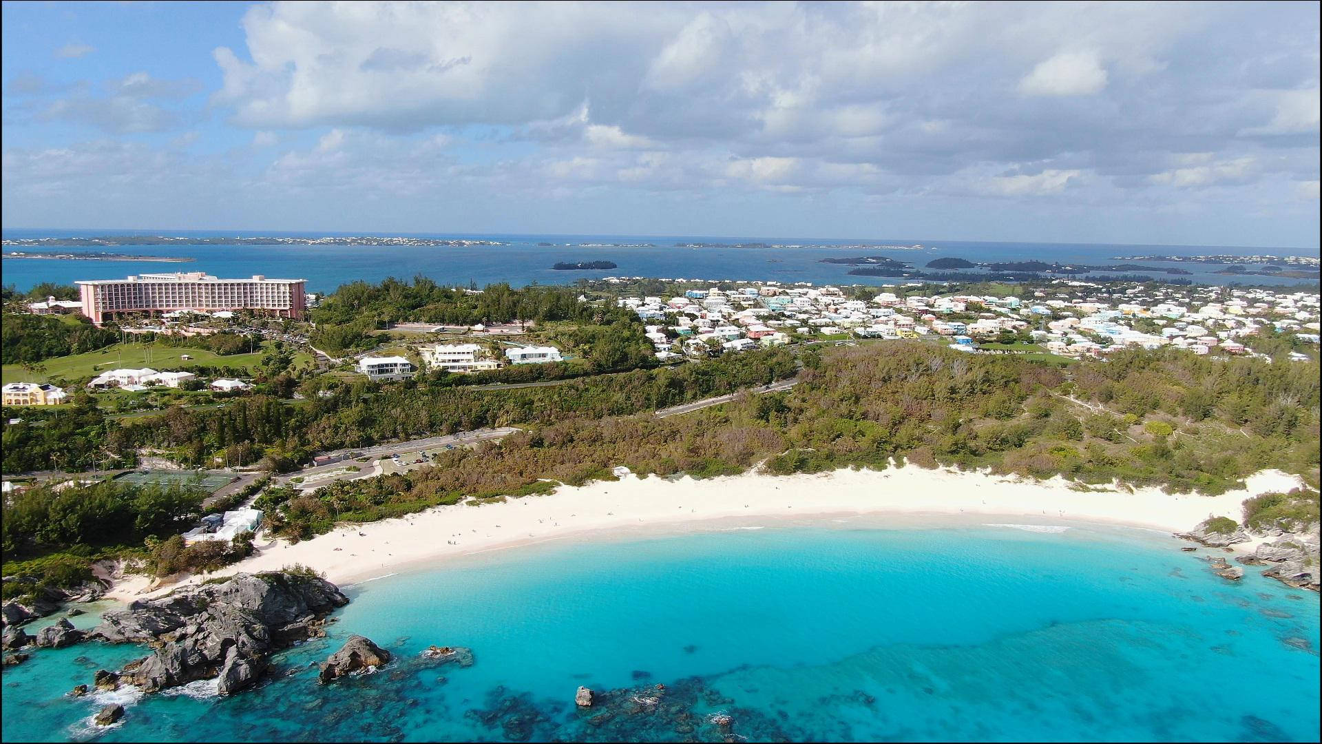 Bermuda 桌布 壁紙 — 新月形藍綠色海灘的鳥瞰圖，白色沙灘被茂密的綠色山丘和度假村建築環繞，天空部分多雲。