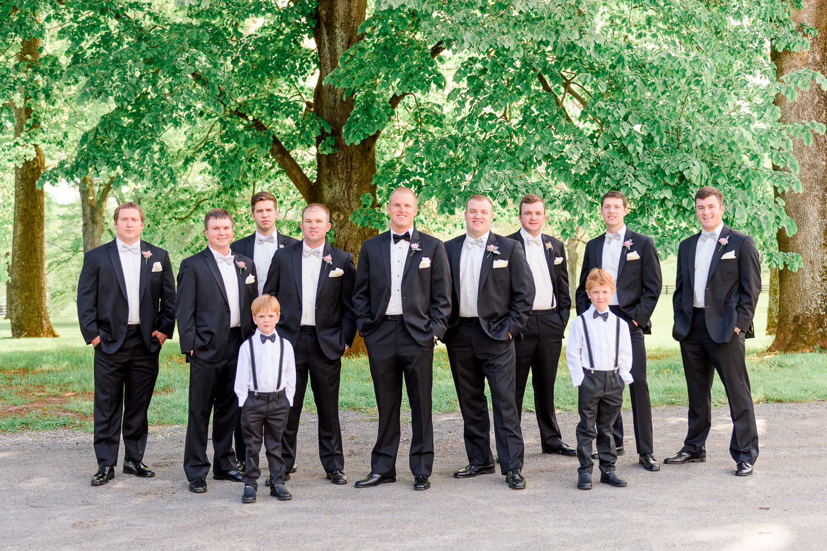 Groomsmen 桌布 壁紙 — 新郎和伴郎們穿著黑色燕尾服，佩戴粉紅色襟花，在陽光明媚的日子裡在茂密的綠樹下擺姿勢。