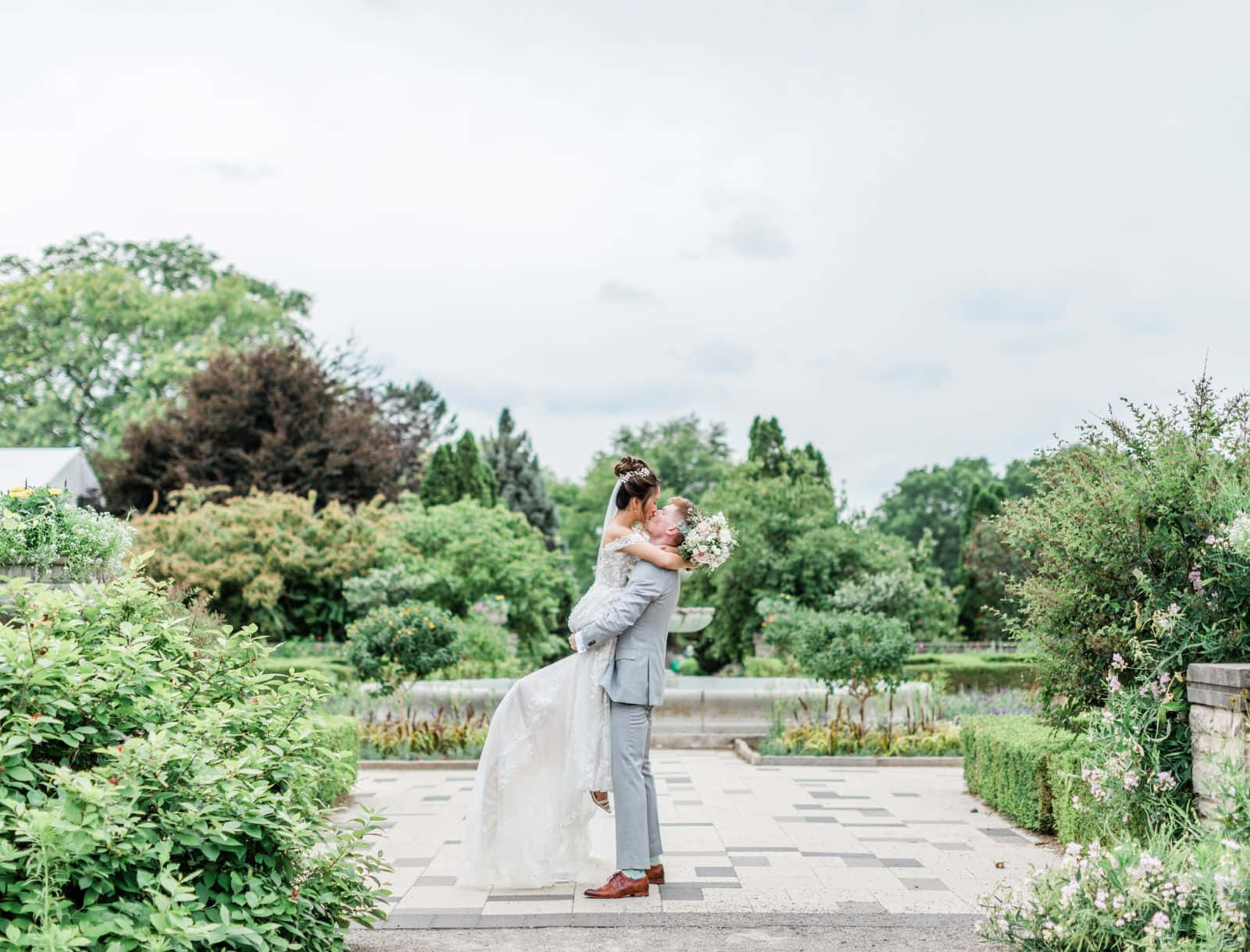 Garden Wedding 桌布 壁紙 — 新郎在茂密的花園中抱起新娘，她手持花束，兩人在多雲的天空下擁抱。