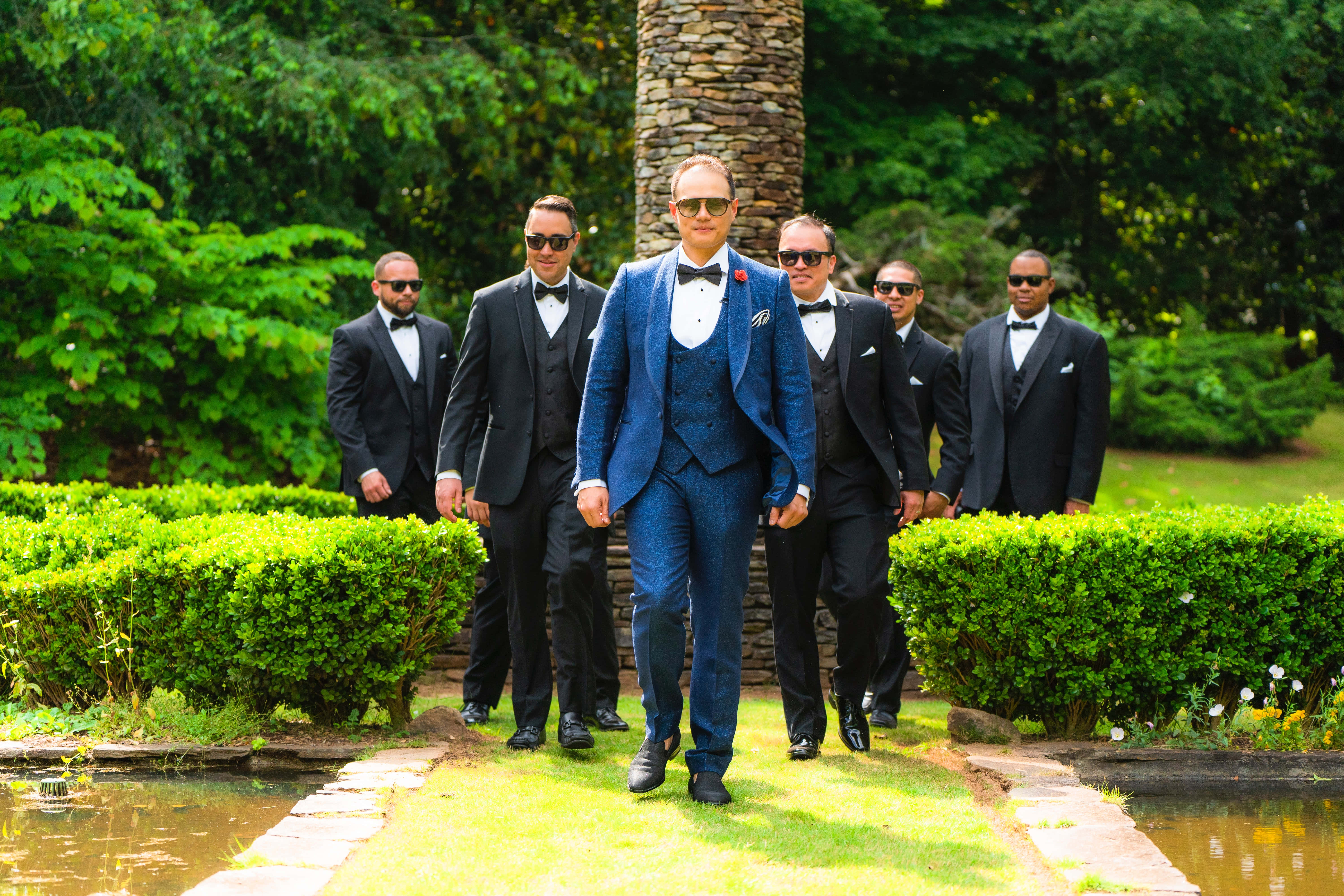 Groomsman 桌布 壁紙 — 新郎穿著鮮藍色西裝，自信地走在花園小徑上，兩旁是穿著黑色燕尾服的伴郎。