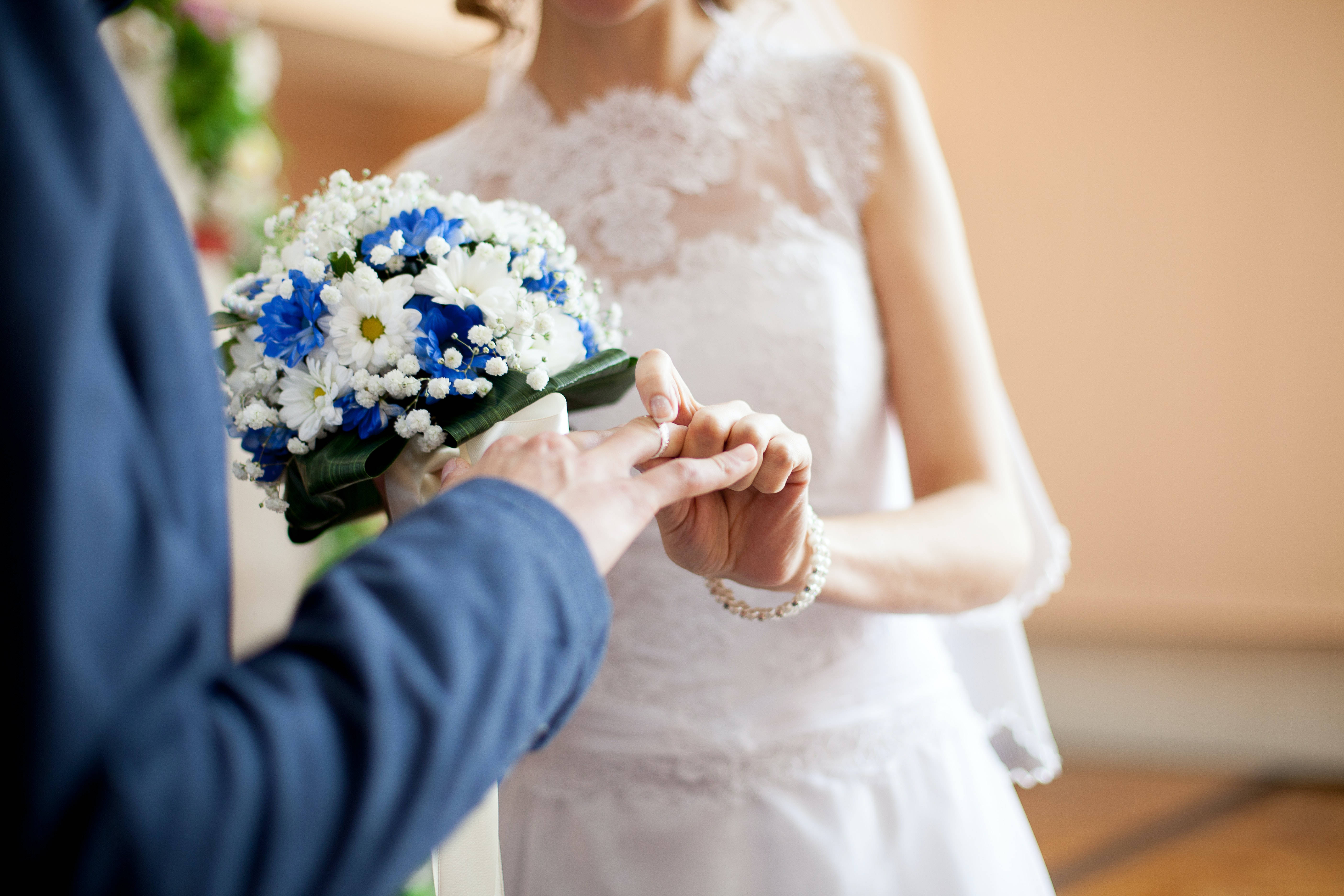 Couple Hands 桌布 壁紙 — 新郎輕輕地將結婚戒指戴在新娘的手指上，新娘手持一束鮮豔的藍白花束。